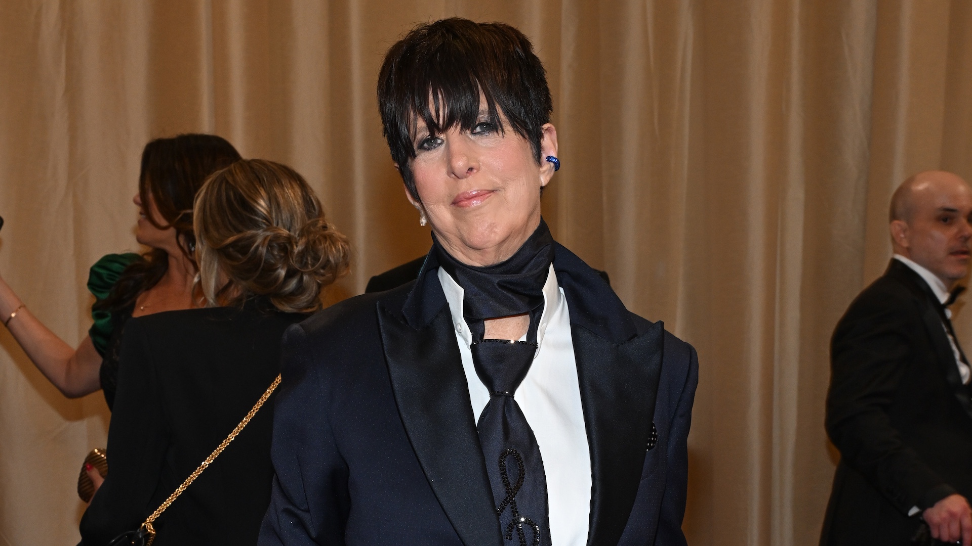 A person with short dark hair wearing a black suit and scarf at an event. Other attendees are in the background.