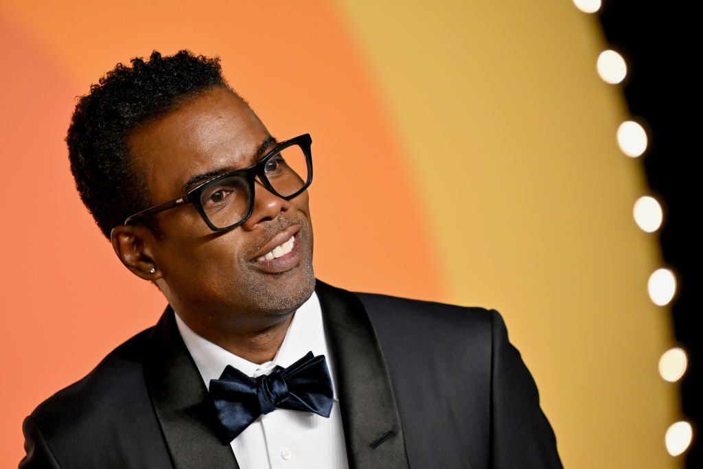 Chris Rock in a tuxedo with a bow tie, wearing glasses, smiling at an event with a bright background.