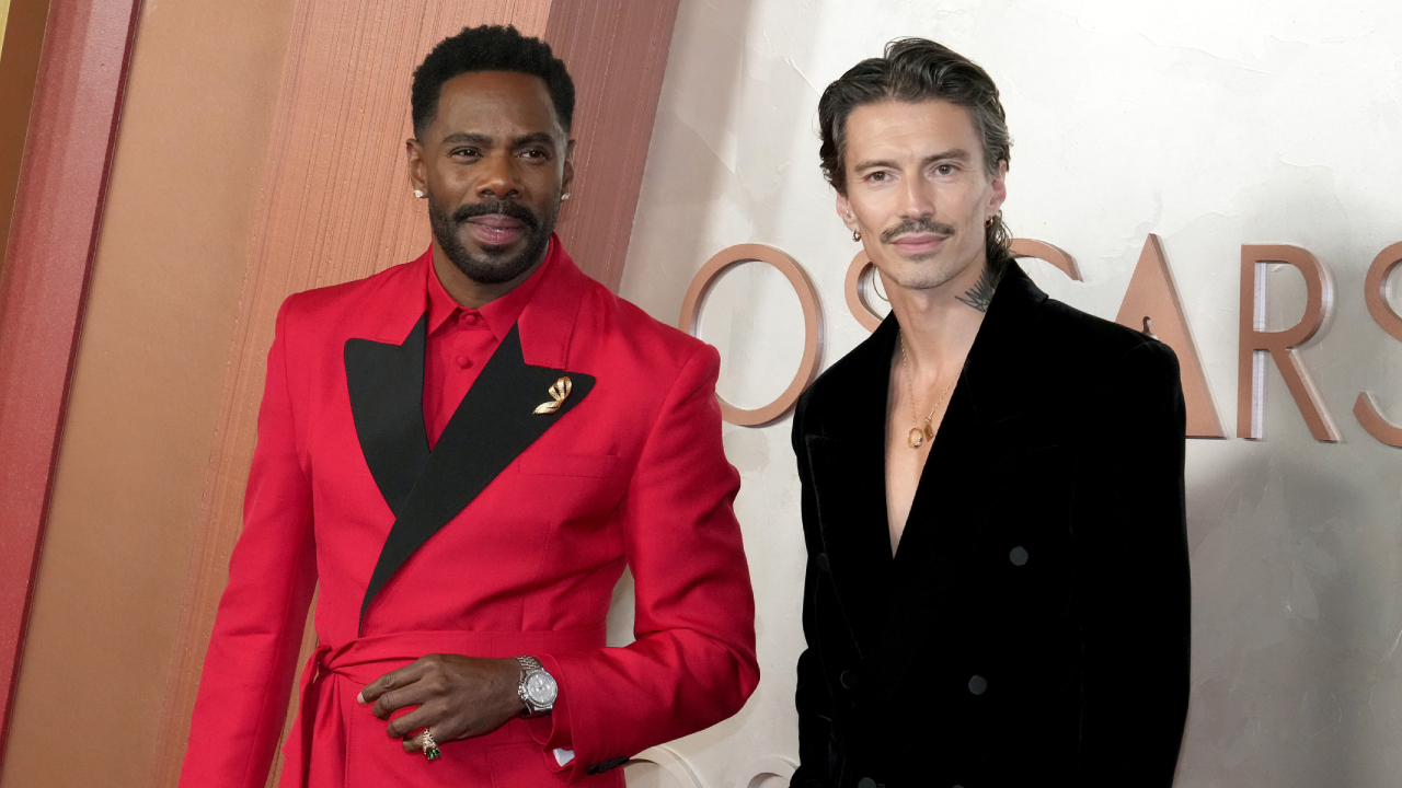 Colman Domingo with husband Raúl in stylish attire at the 2025 Oscars. Colman wears a red Valentino suit with black accents, Raúl a black suit with a gold necklace.