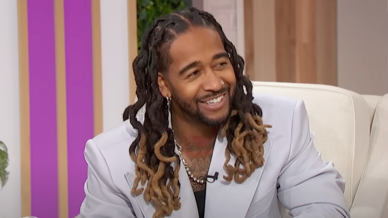 Omarion with long dreadlocks and a light gray suit is smiling while sitting on a couch in a studio setting.
