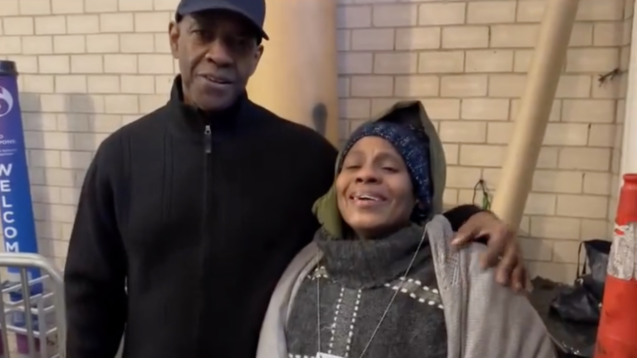A man in a black jacket and cap stands with his arm around a smiling woman in a gray sweater and hat, in front of a brick wall.