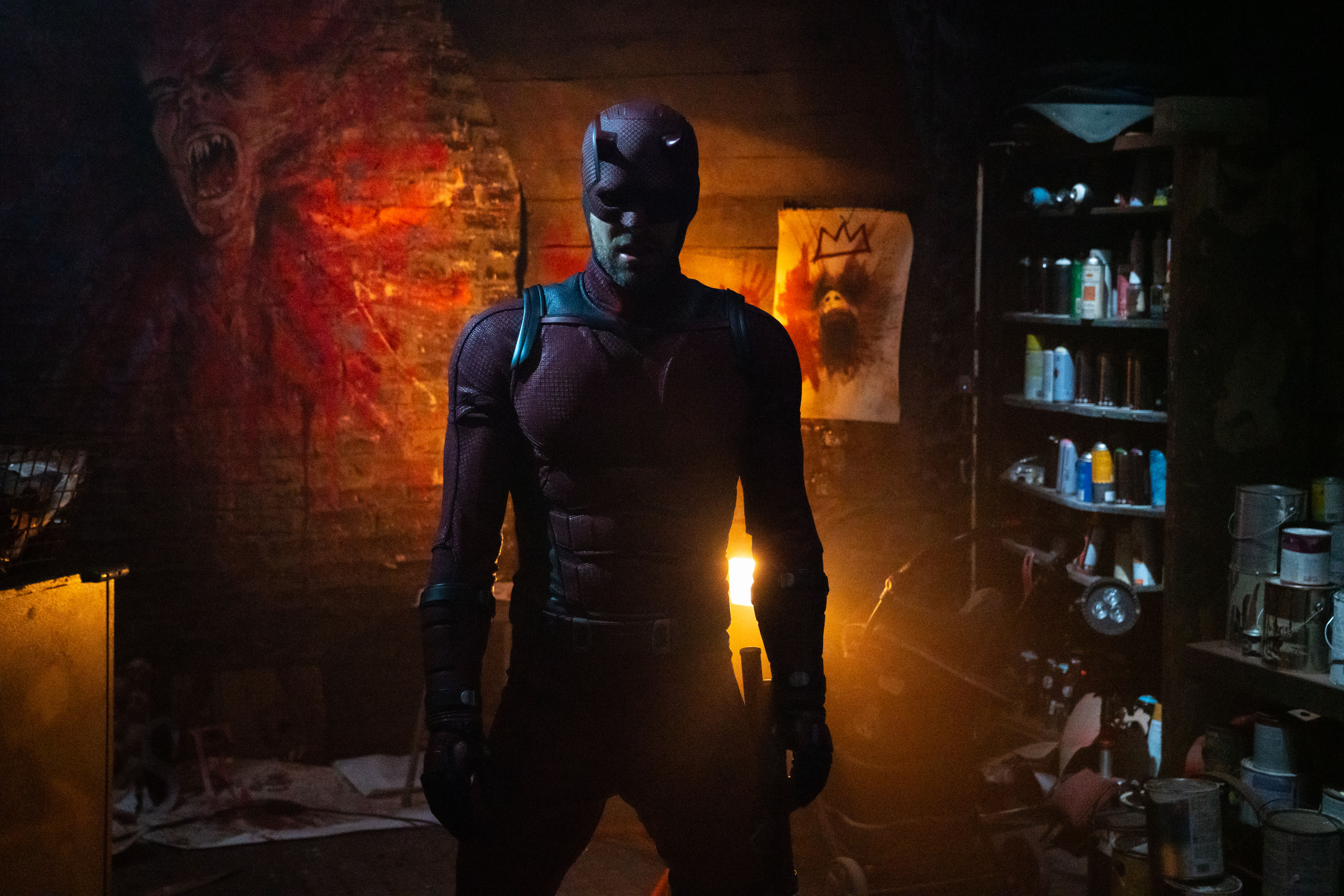 A person in a Daredevil costume stands in a dimly lit room with graffiti on the walls and shelves filled with paint cans.