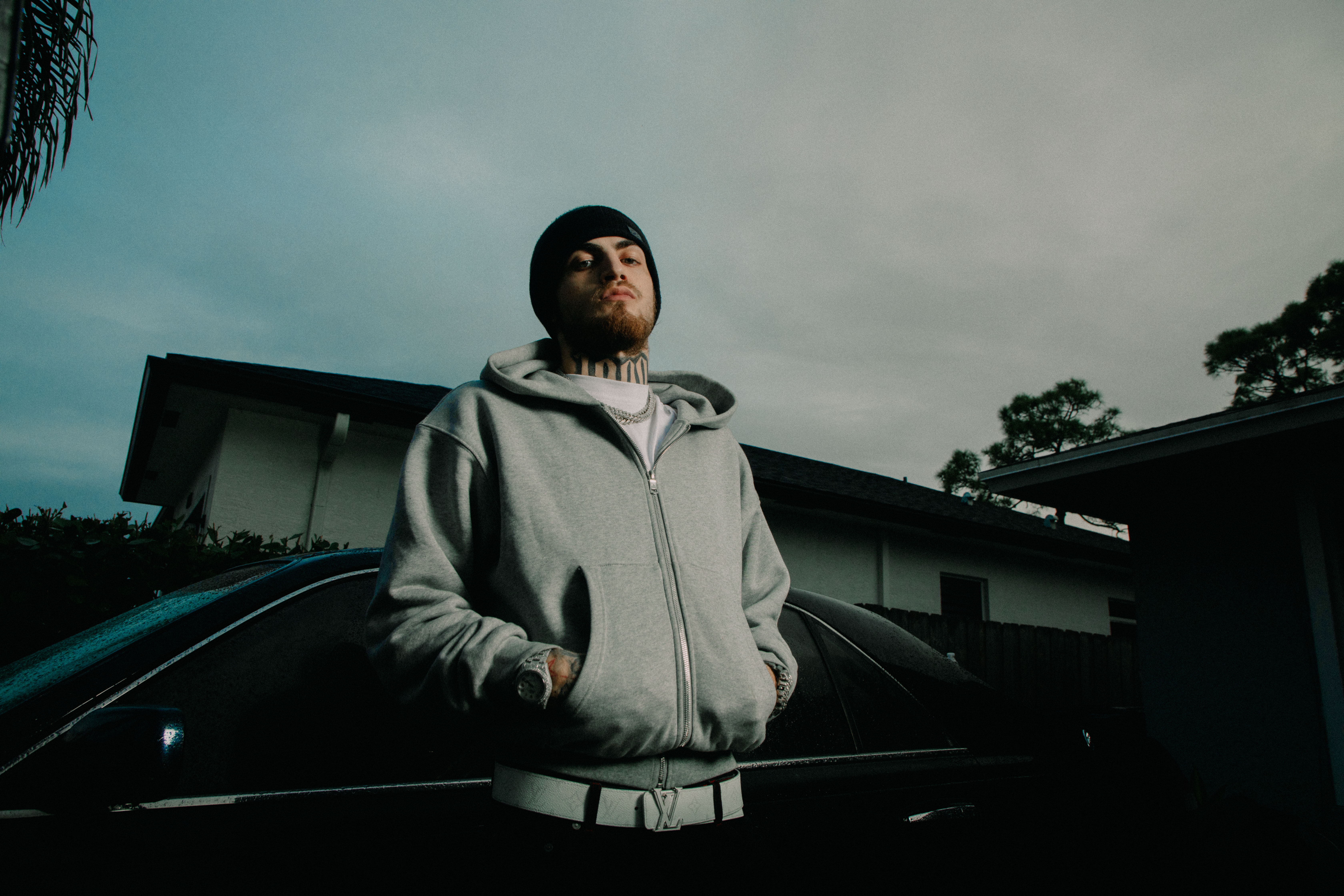 1900Rugrat in a gray hoodie and beanie stands in front of a car, with a house and trees in the background, under a cloudy sky.