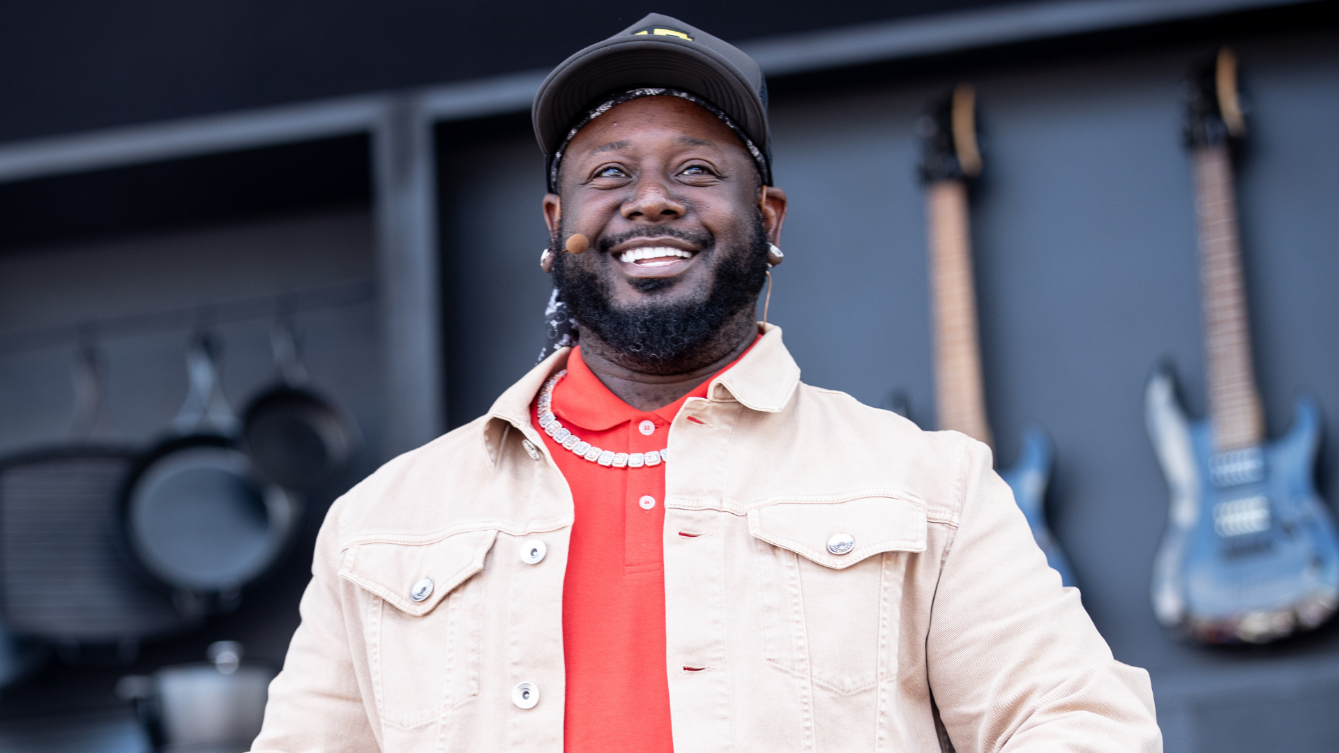 T-Pain smiling, wearing a beige jacket and red shirt, with guitars in the background.