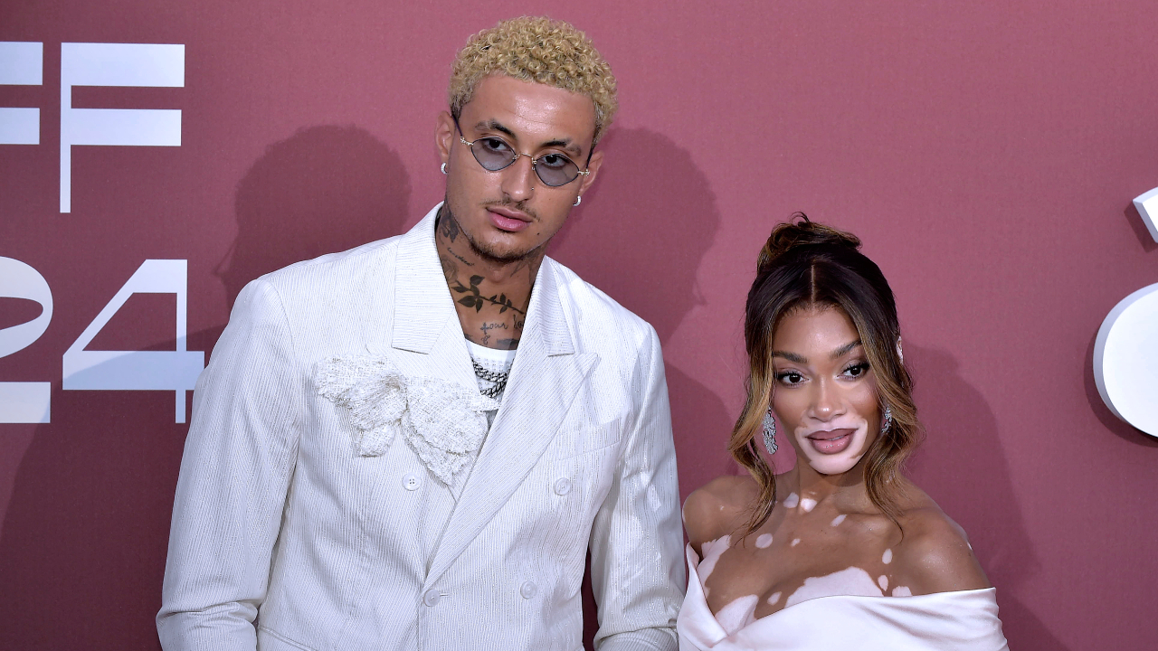 Kyle Kuzma in a white suit and Winnie Harlow in an elegant white dress pose together at a red carpet event.