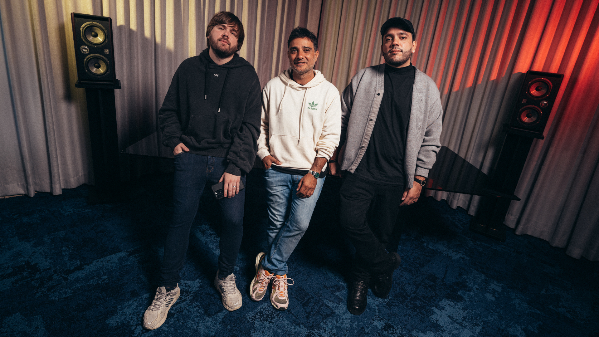 Three men standing in a room with speakers, wearing casual clothing. The background features curtains with a warm lighting effect.