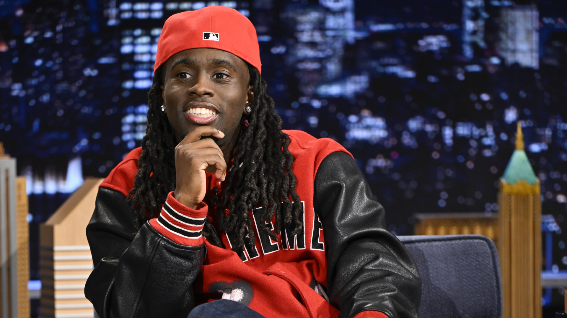 A person wearing a red cap and jacket with "Supreme" sits on a talk show set, smiling with a cityscape background.
