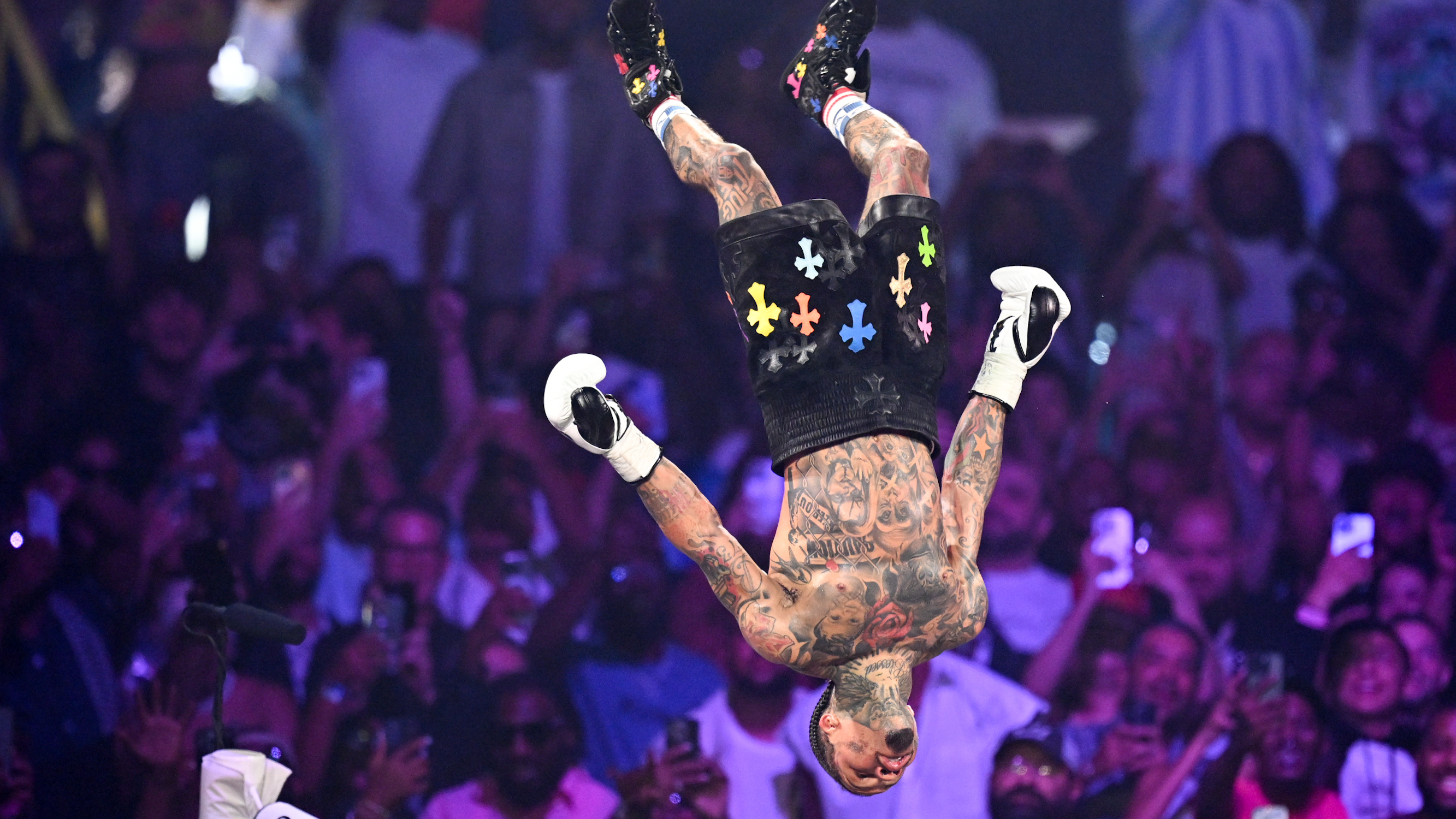 A performer doing a backflip on stage in front of a cheering crowd. The person is tattooed and wearing black shorts with colorful designs.