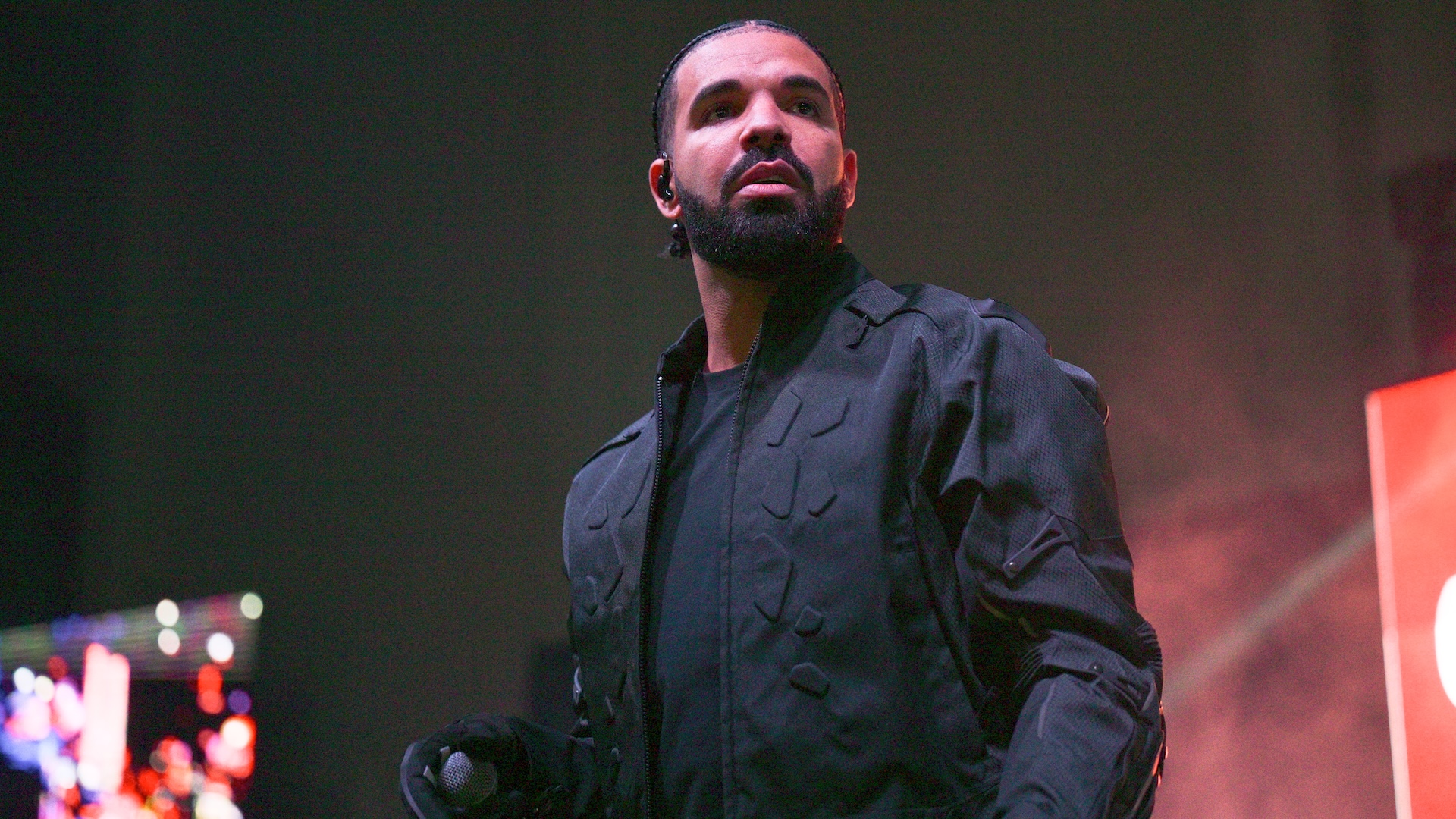 Drake performing on stage, wearing a black jacket, holding a microphone, with a focused expression and stage lights in the background.