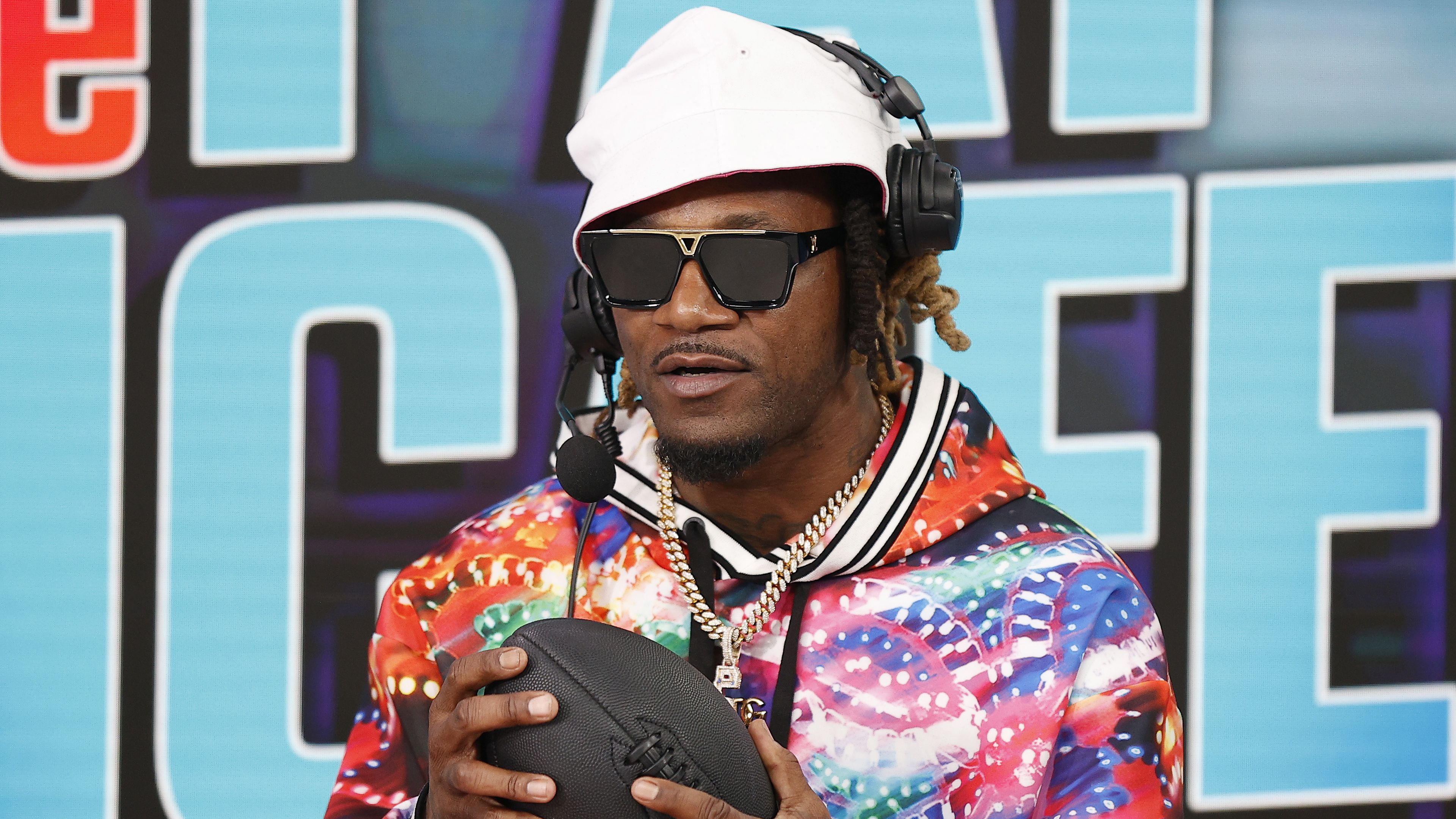 Adam "Pacman" Jones in colorful attire and sunglasses, wearing a headset, holds a football.