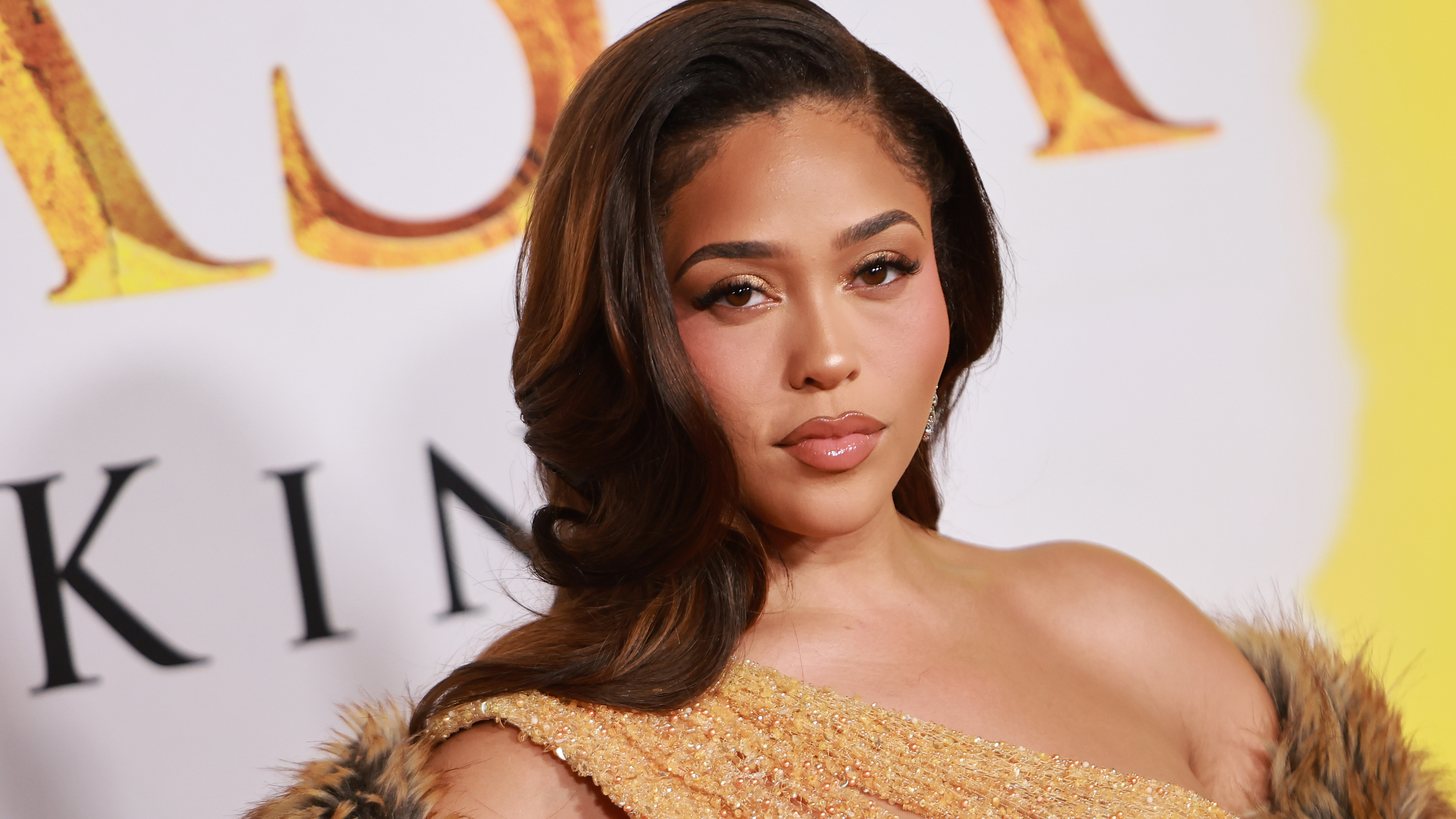 Jordyn Woods in a glamorous gold dress with a fur coat poses on a red carpet with a yellow and white background.