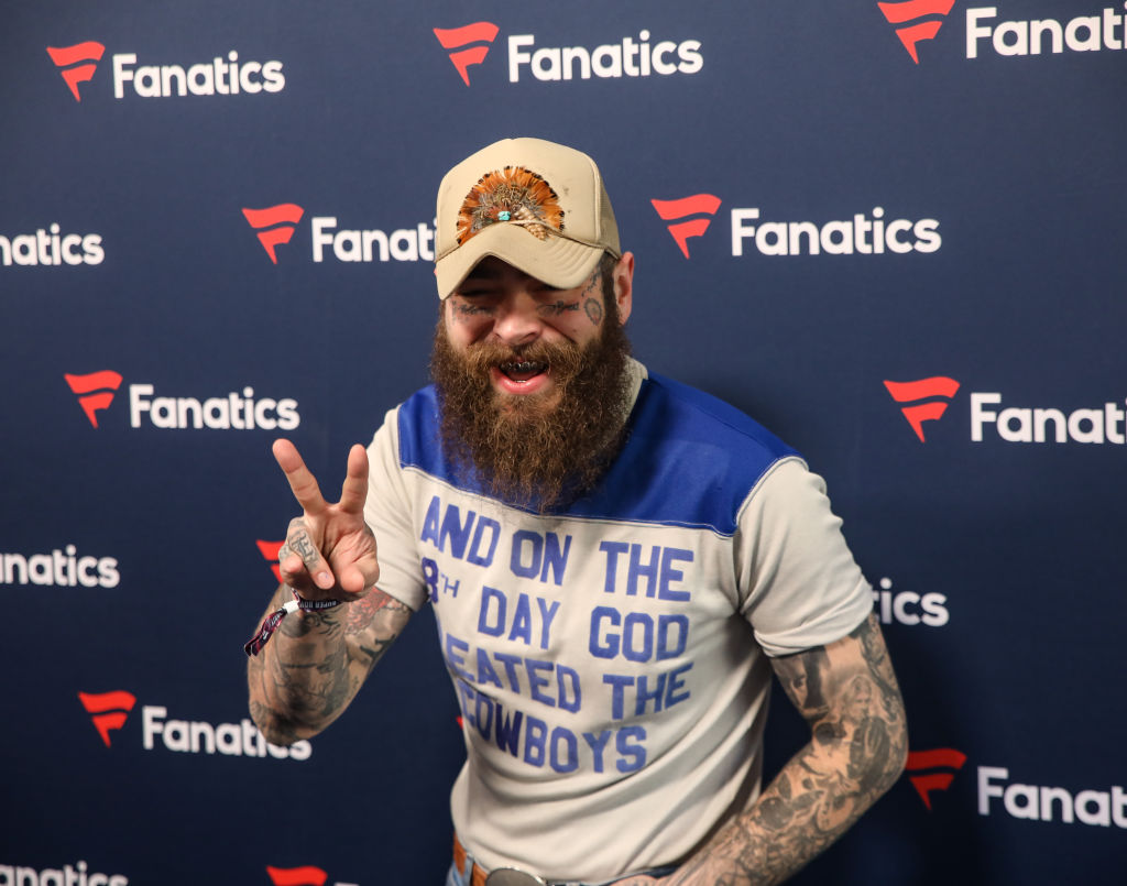A man with a beard and tattoos wearing a cap and a shirt with text poses in front of a Fanatics backdrop, making a peace sign.
