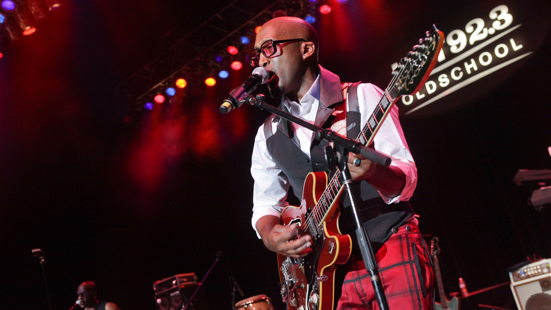 A musician in red plaid pants and glasses plays guitar and sings on stage under colorful lights, with a "92.3 Old School" sign.