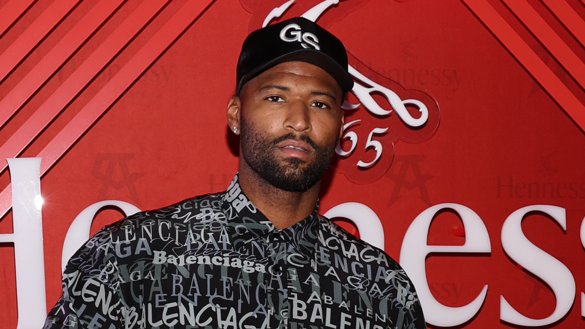 A man wearing a Balenciaga shirt and a black cap stands in front of a red Hennessy backdrop.
