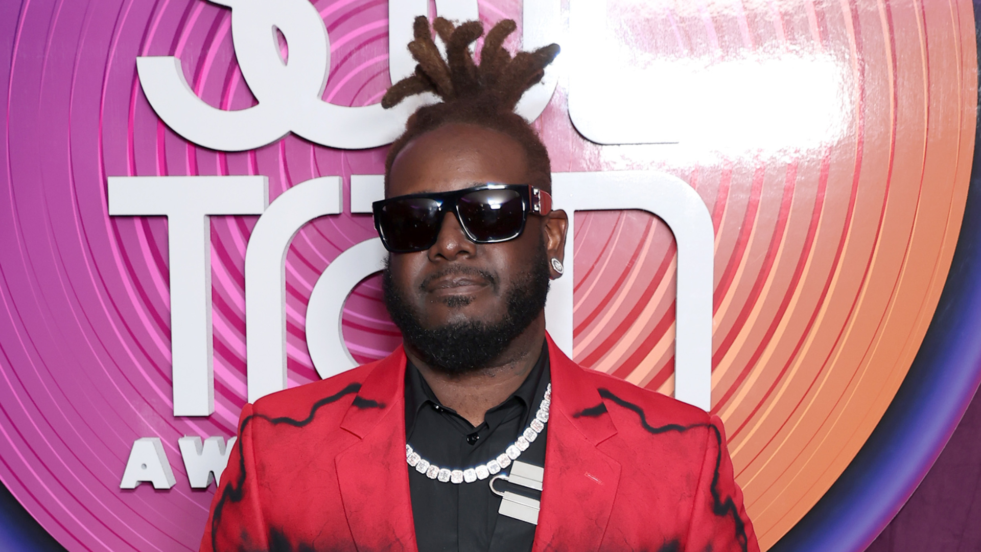 T-Pain wearing sunglasses and a red suit, standing in front of a colorful backdrop with "Soul Train Awards" text.