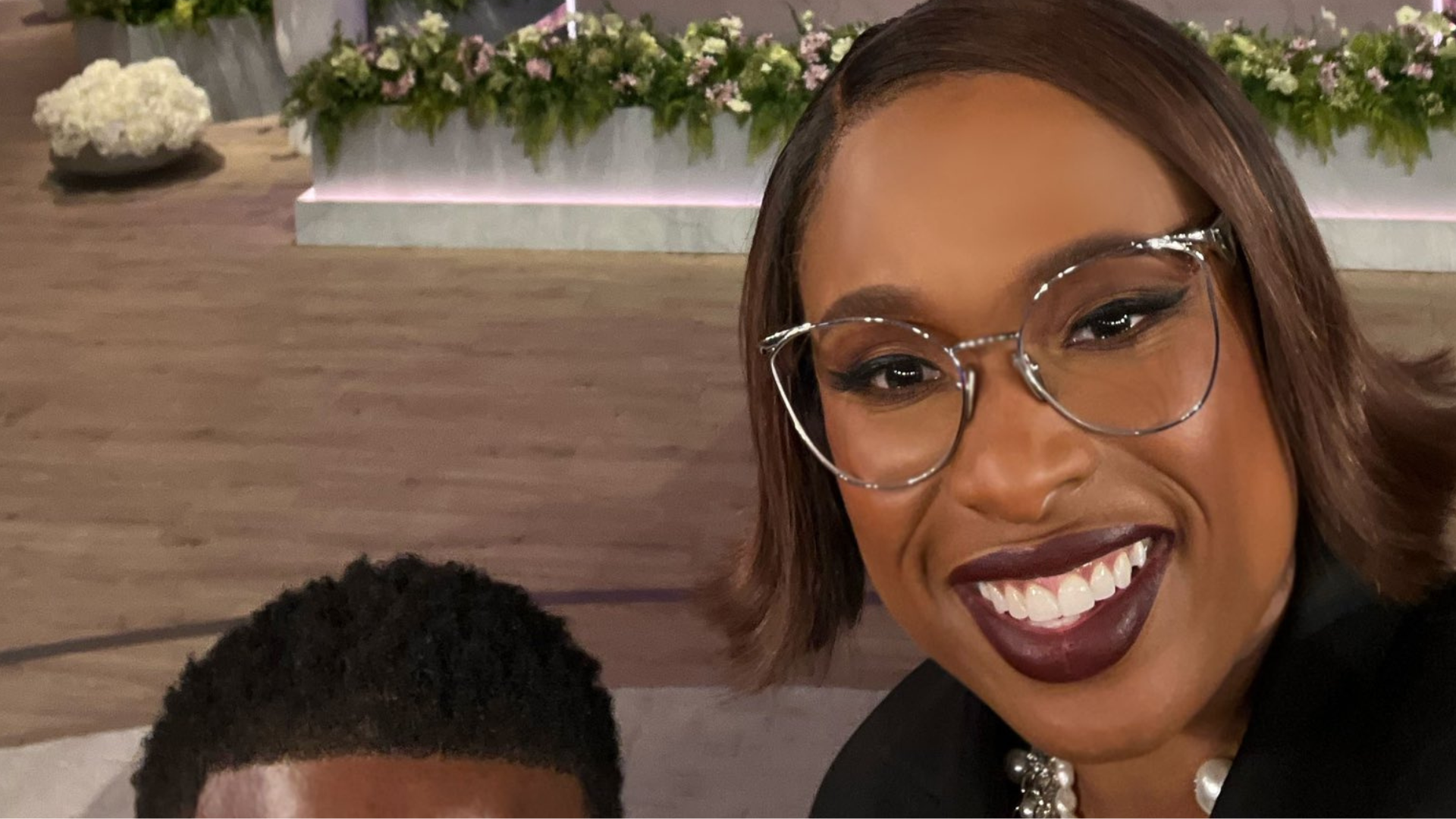 A woman smiling with glasses and dark lipstick, taking a selfie indoors with floral decorations in the background.