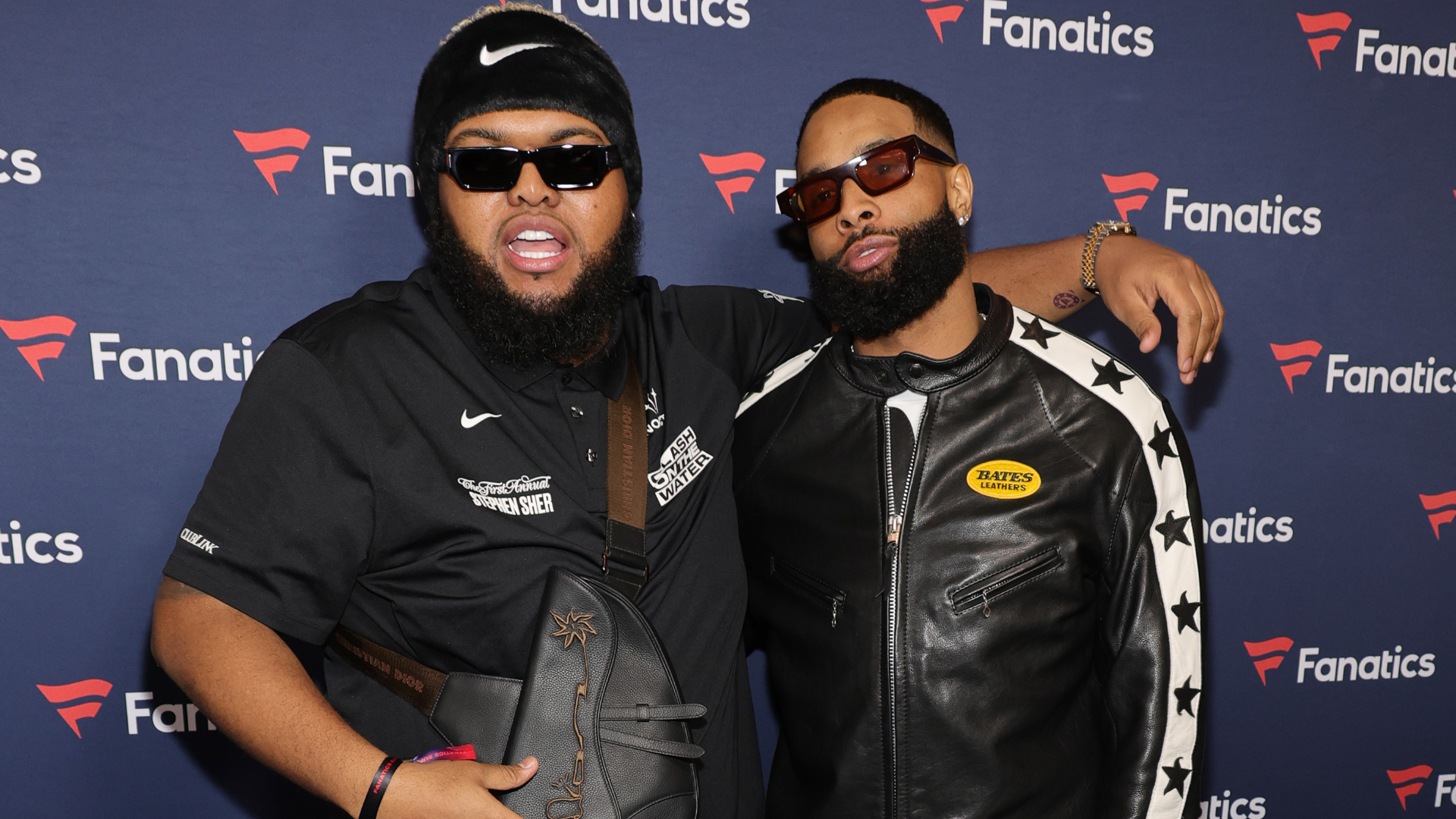 Two men wearing sunglasses and stylish outfits pose together at a Fanatics event, with a branded backdrop behind them.