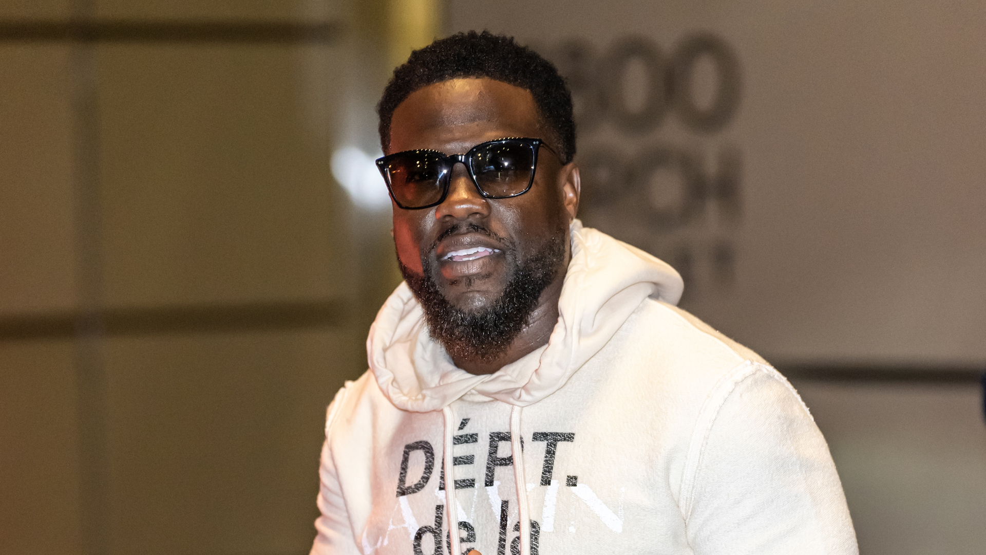 Kevin Hart wearing sunglasses and a light-colored hoodie with text, smiling in a casual setting.