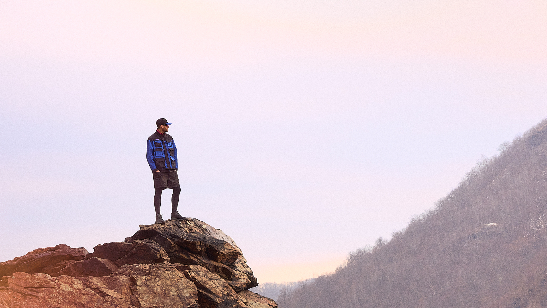 A person in outdoor gear stands on a rocky cliff, overlooking a misty valley. The sky is soft pink and orange.