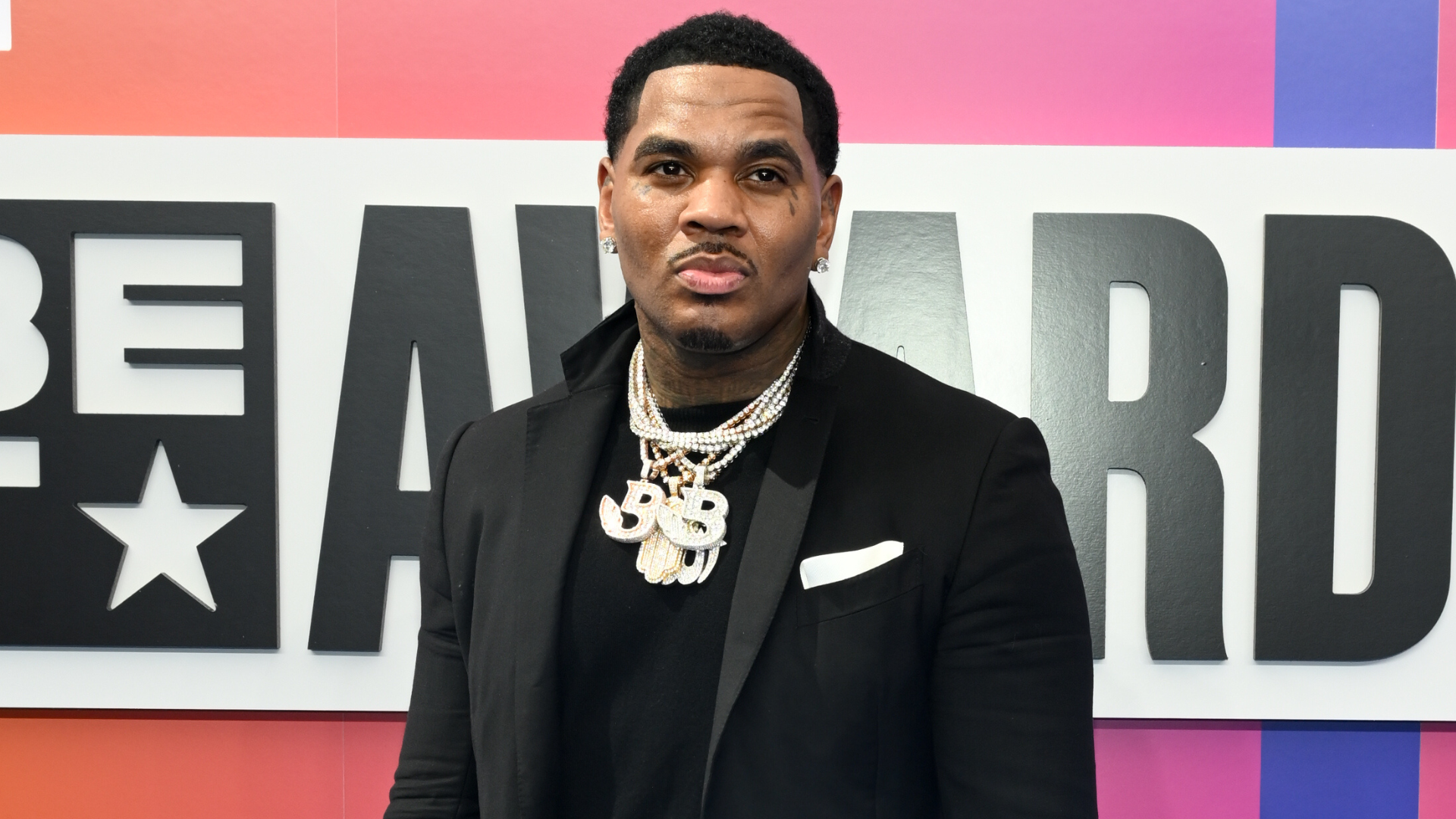 A man in a black suit with layered necklaces stands in front of a BET Awards sign.