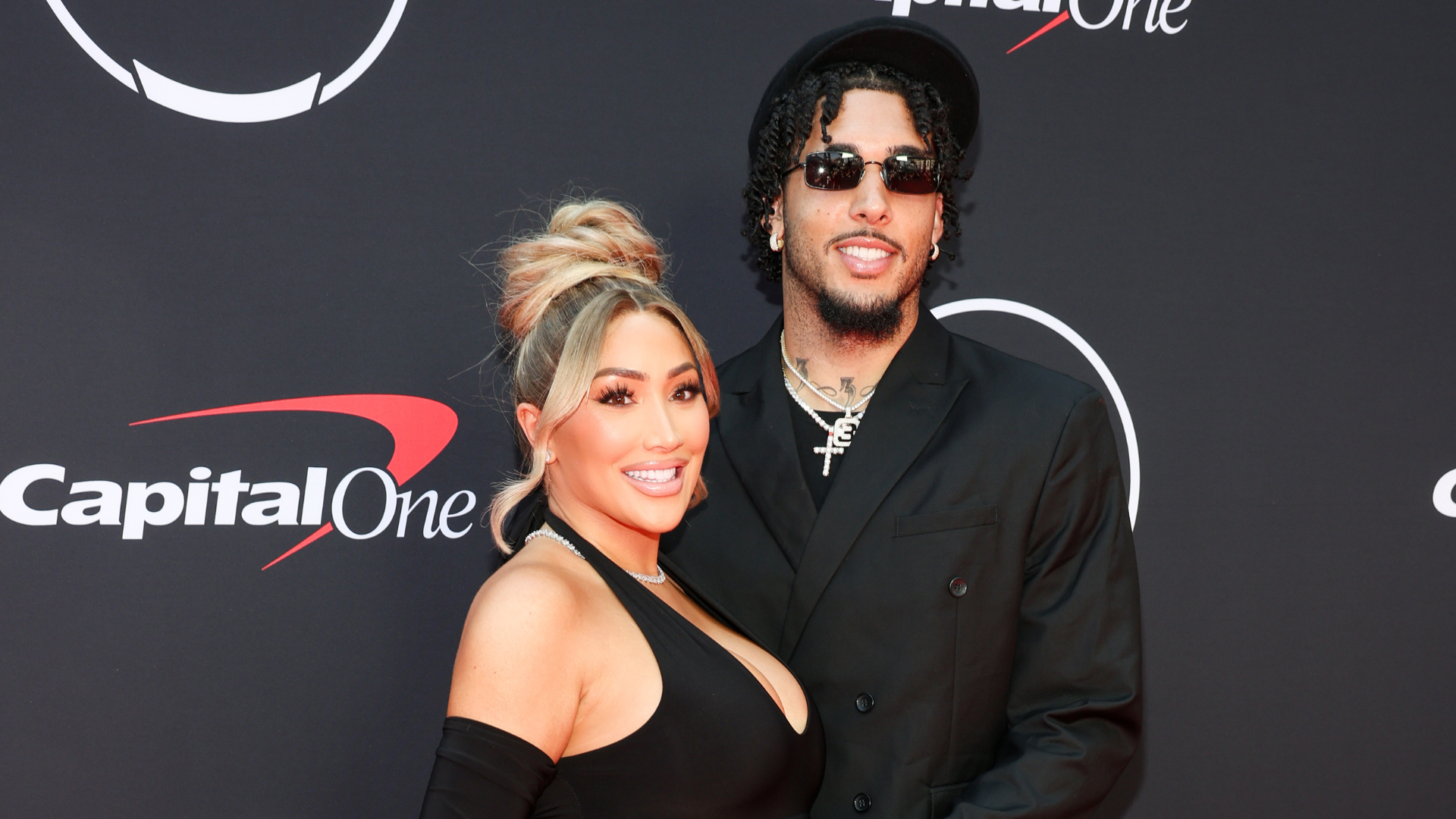 A couple poses on the red carpet. The woman is in a black dress, and the man is in a black suit with sunglasses.