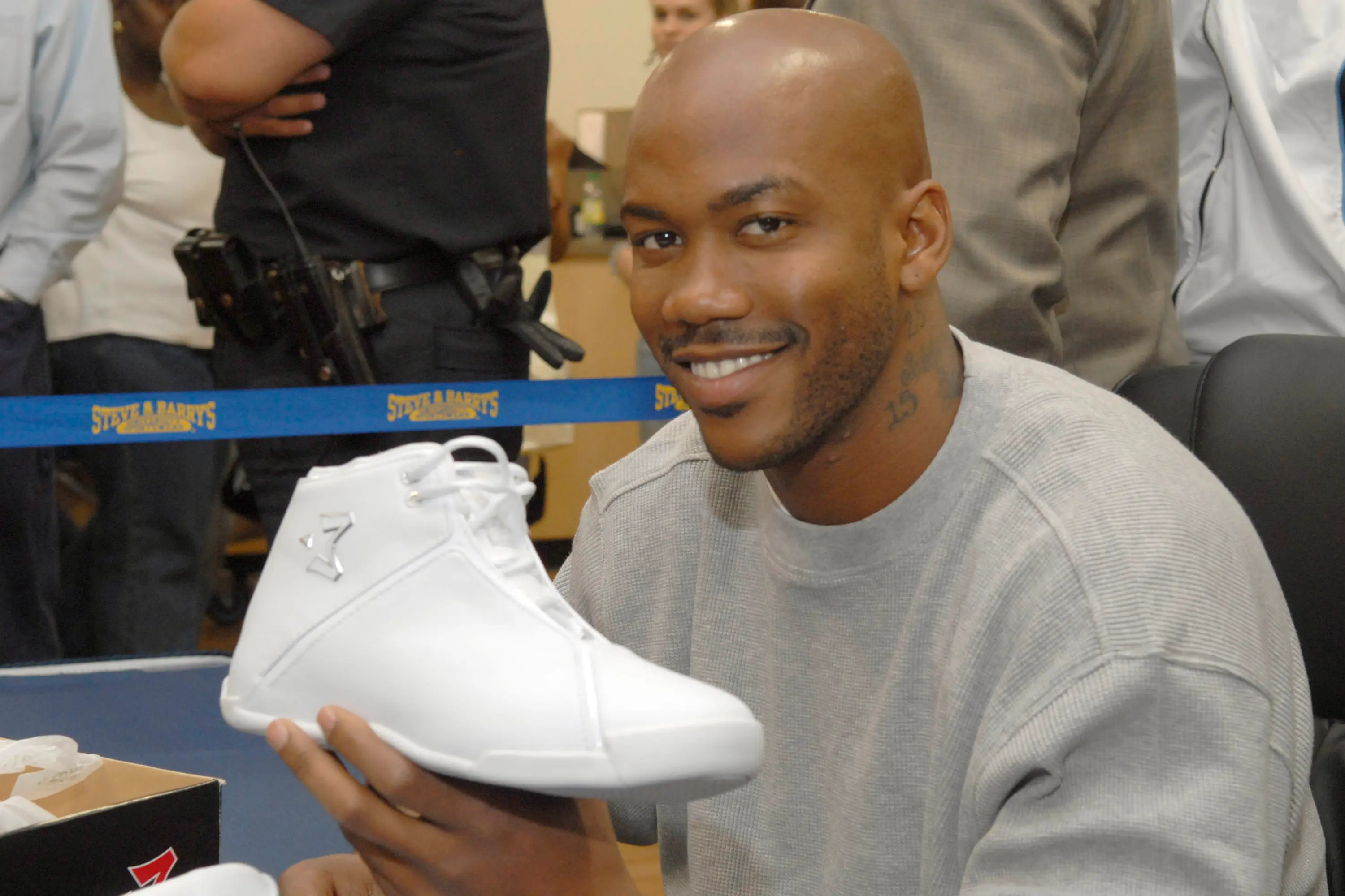 A person holding a white sneaker at a public event, smiling, with a crowd and security in the background.