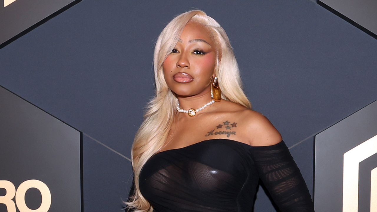 A woman with long blonde hair and a black off-shoulder top poses in front of a patterned background.