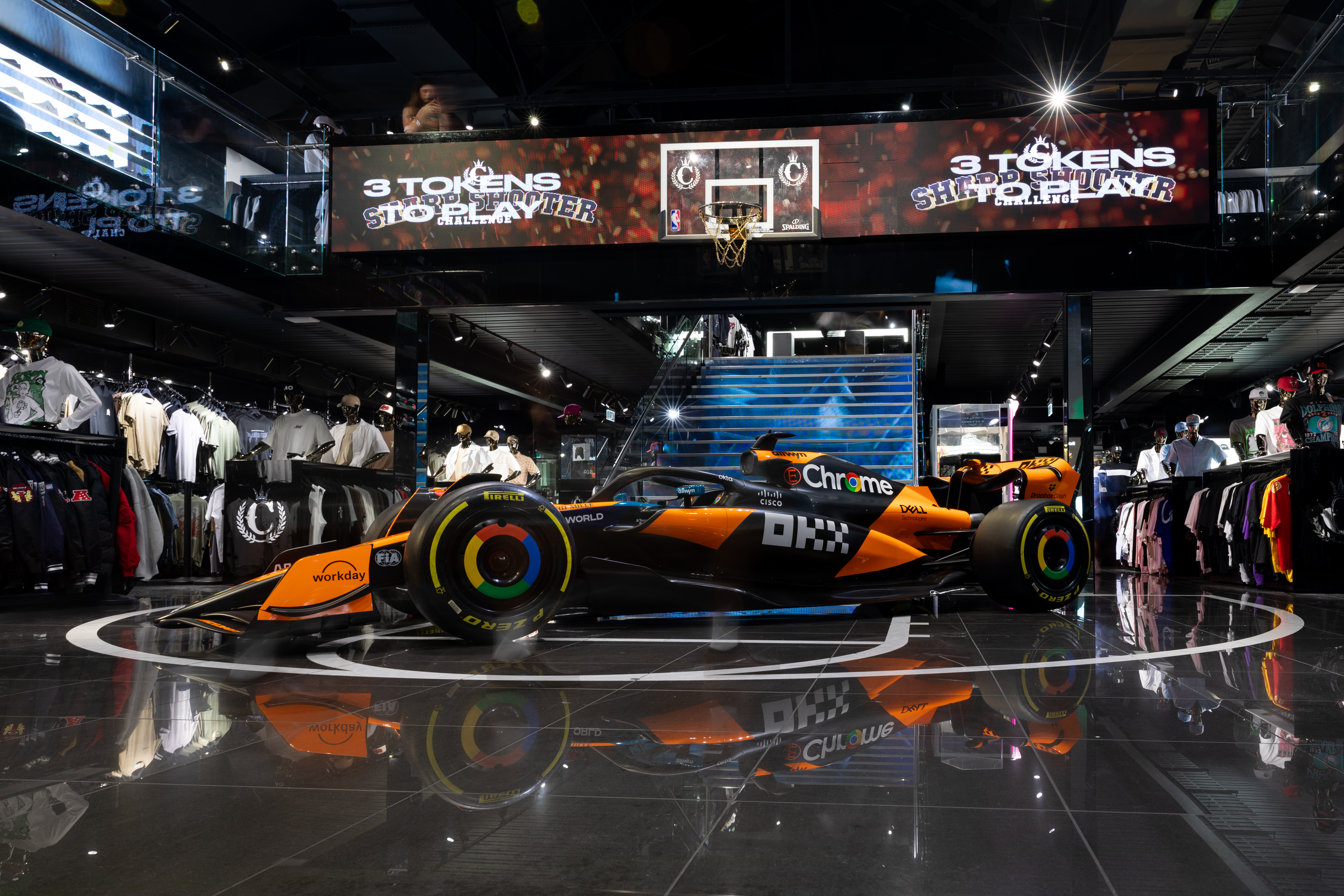 A Formula 1 car displayed in a clothing store with merchandise around. A basketball hoop is above, and digital screens display text.