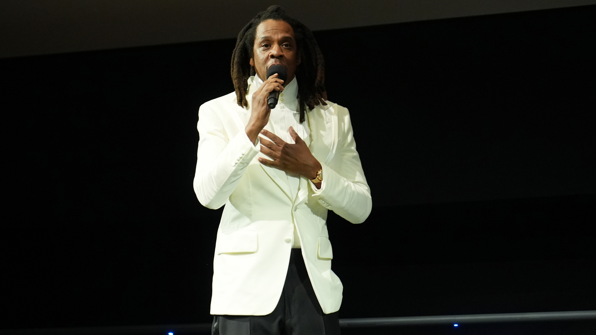 A man in a white suit with dreadlocks holds a microphone on stage.