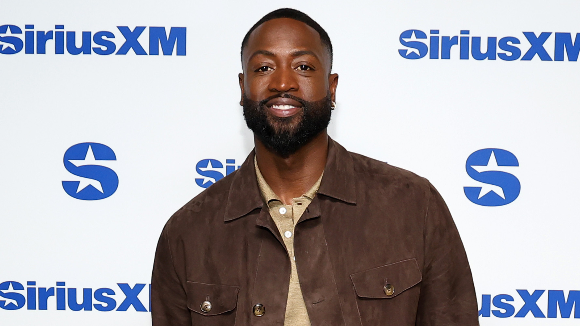 Dwyane Wade at a SiriusXM event, wearing a brown jacket over a beige shirt, smiling against a branded backdrop.