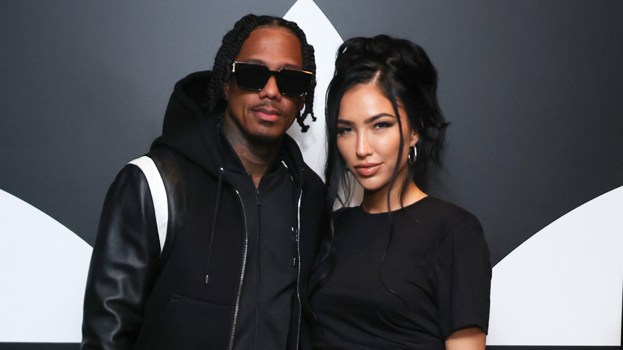 Nick Cannon in sunglasses and Bre Tiesi with dark hair pose together in front of a black and white background.