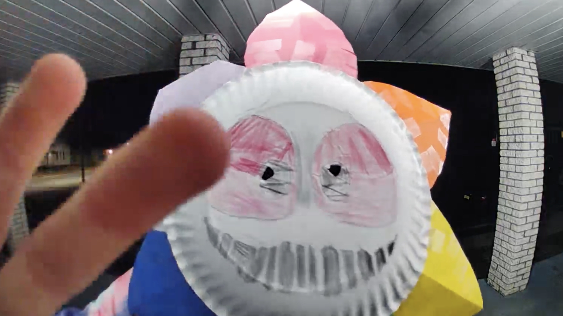 Person in a colorful costume with a paper plate mask featuring a drawn face, making a peace sign gesture towards the camera.