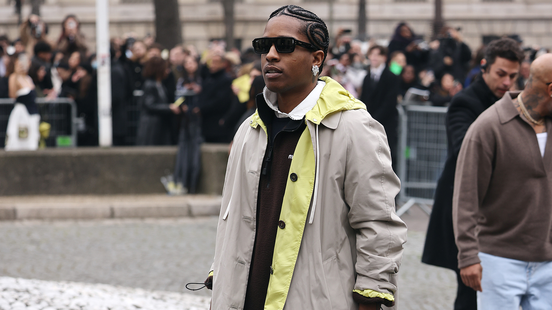 A$AP Rocky wearing sunglasses and a stylish coat, walking on a cobblestone street with a crowd in the background.
