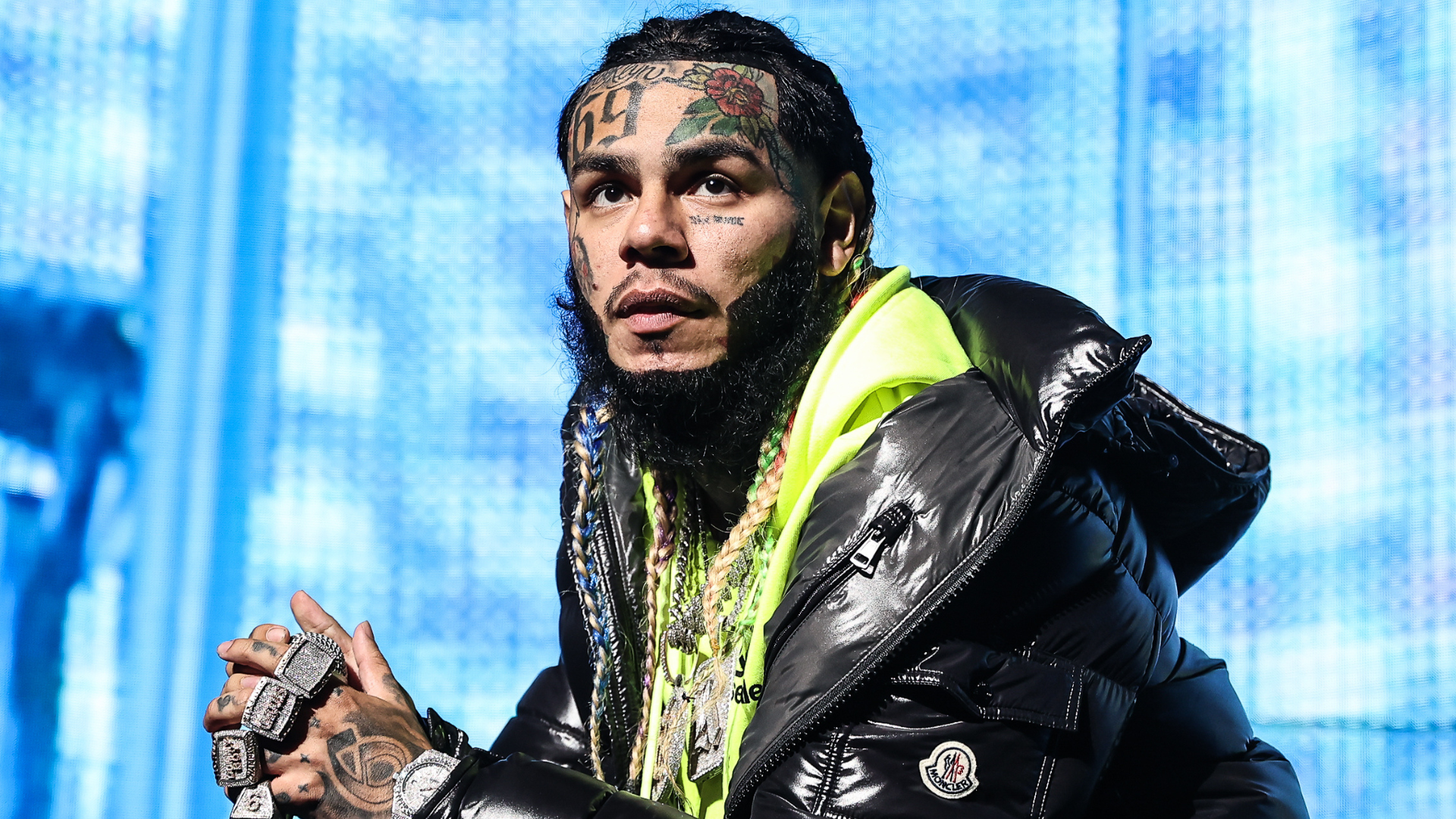 Rapper Tekashi 6ix9ine, with face tattoos and colorful braided hair, wearing a black puffer jacket and neon yellow hoodie.