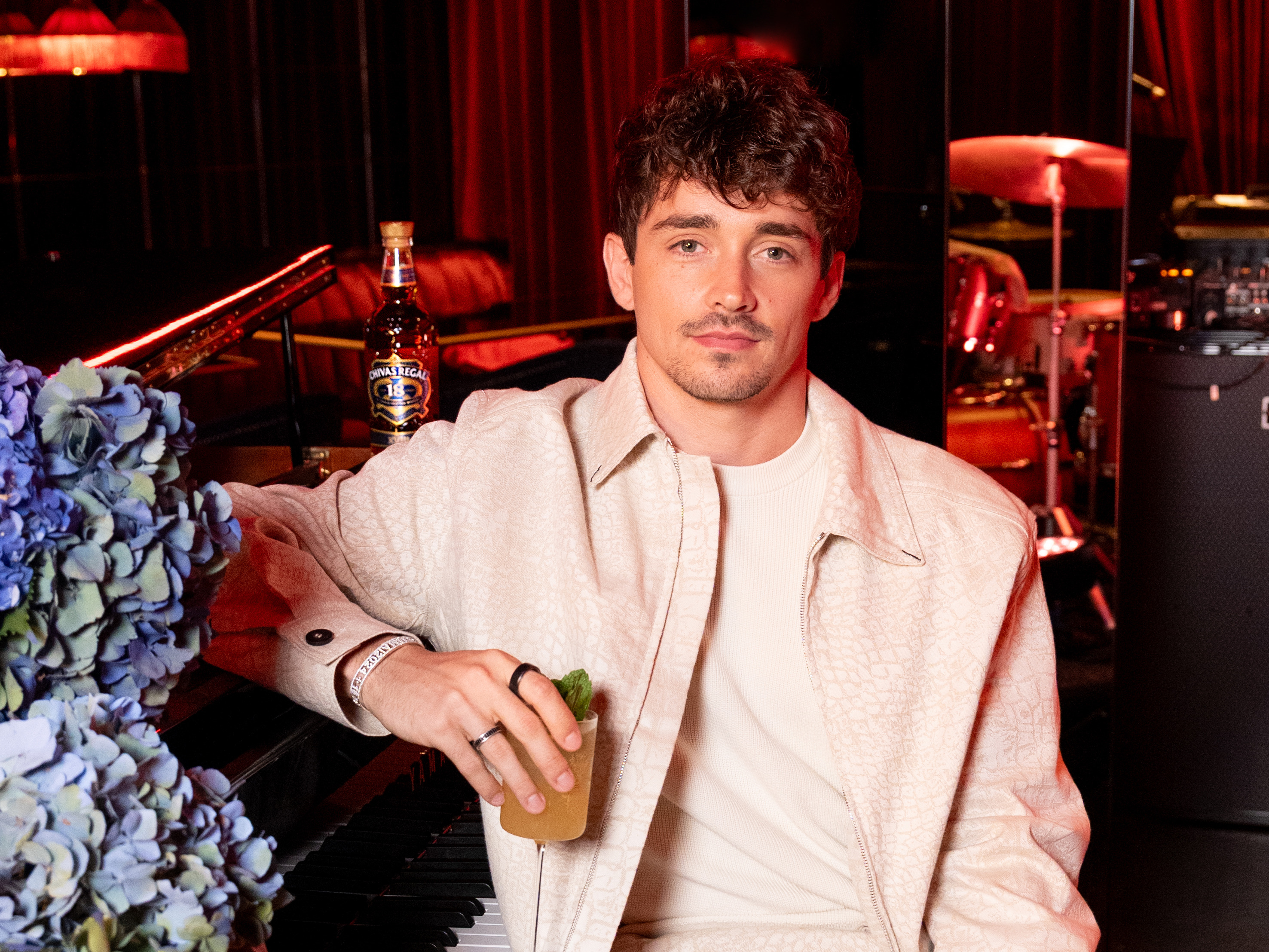 A man in a light-colored outfit sits at a piano holding a drink, with a bottle of Chivas Regal nearby in a dimly lit room.
