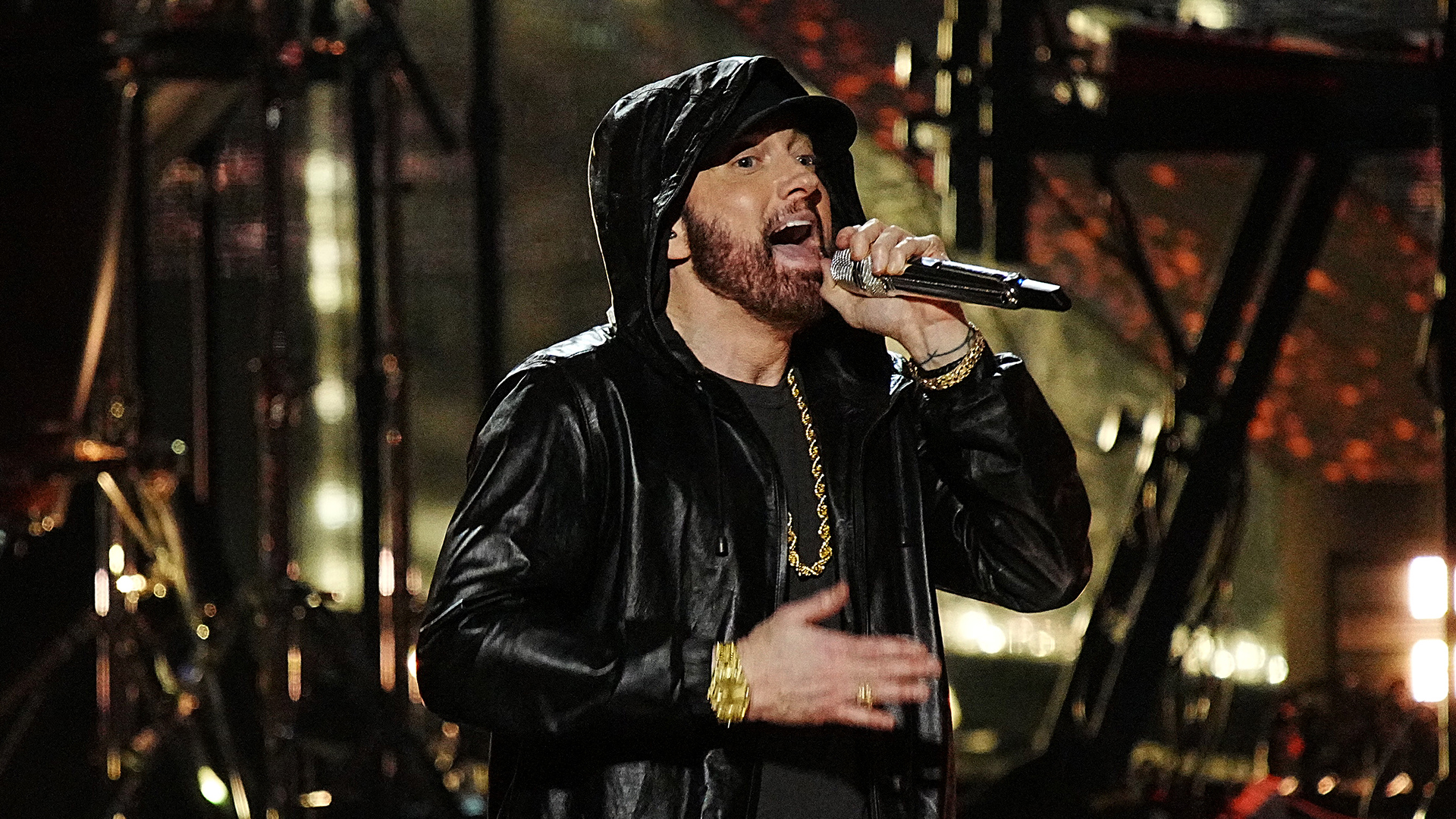 Eminem performing on stage, wearing a black hoodie and jacket, holding a microphone, with a dynamic background.