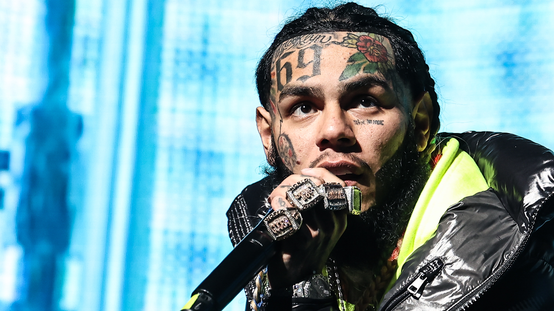 Rapper Tekashi 6ix9ine performing on stage, holding a microphone, wearing a black jacket with visible facial tattoos.