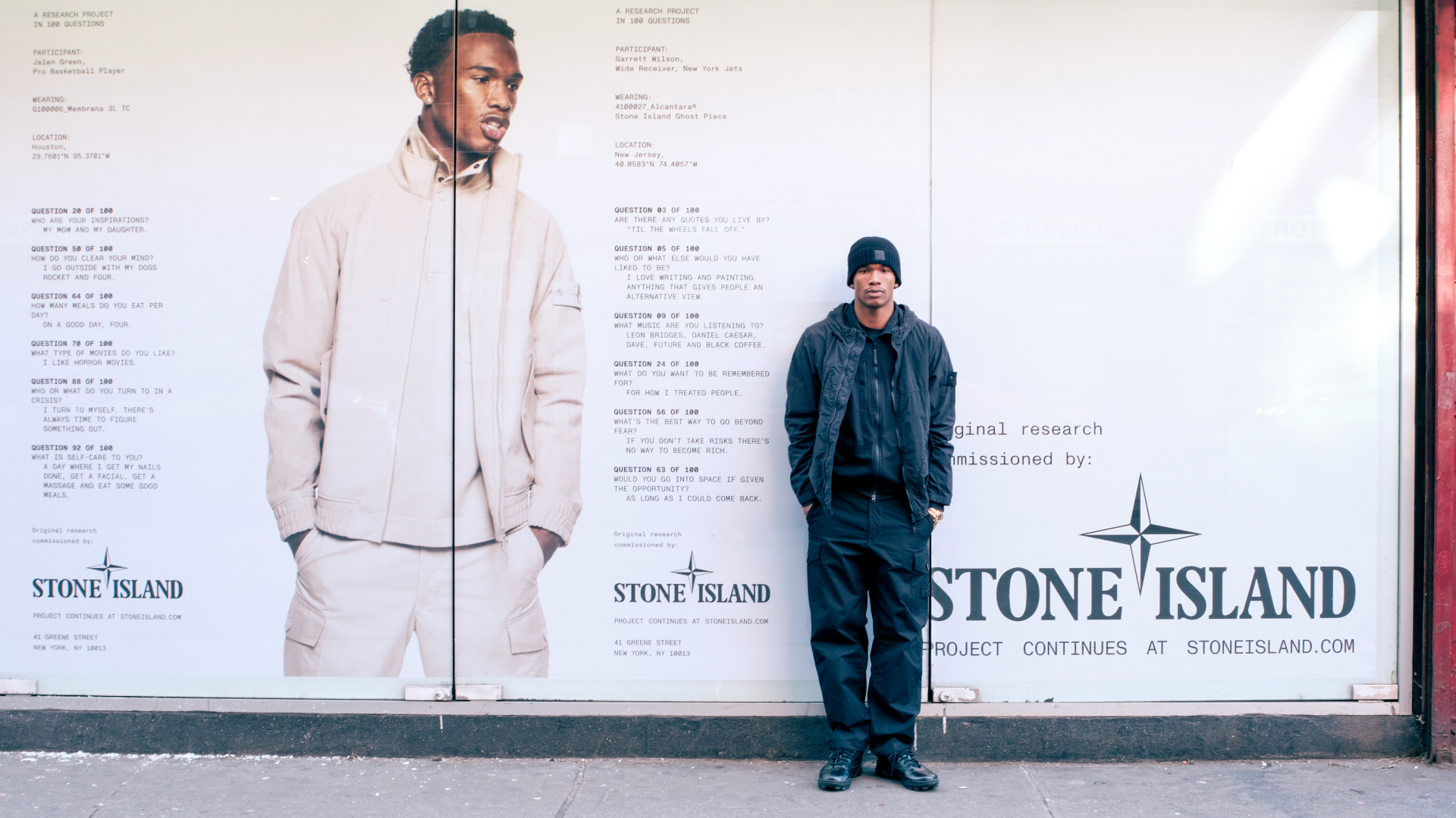 A person stands in front of a large Stone Island advertisement featuring a model in a light-colored jacket, on a city sidewalk.