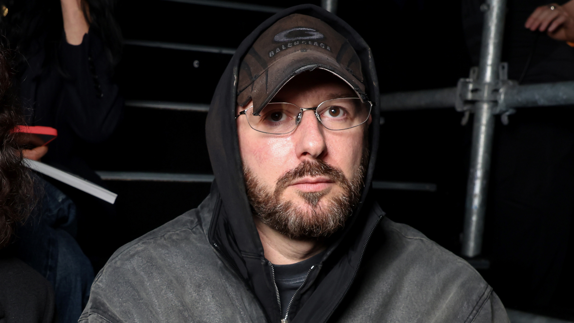 A man with glasses, a beard, and a hooded jacket, wearing a Balenciaga cap, sitting in a dimly lit area.
