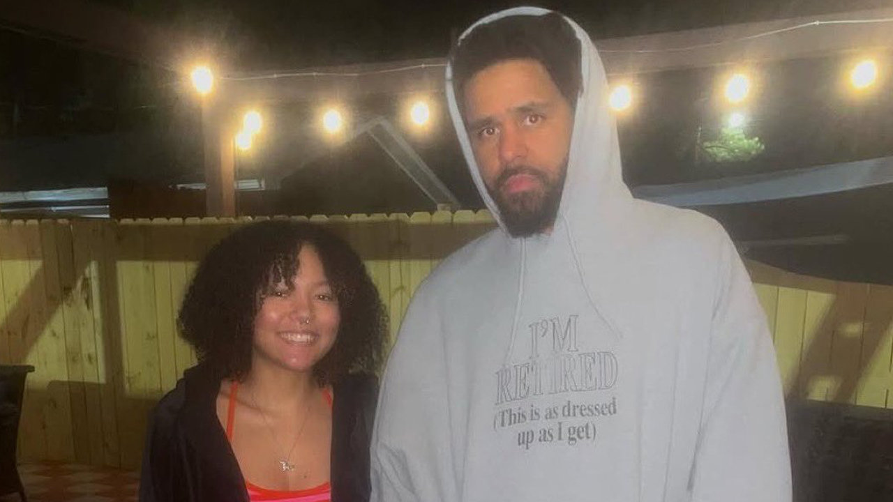 J. Cole posing with a fan while wearing a hoodie that reads, "I'm Retired (This is as dressed up as I get)."
