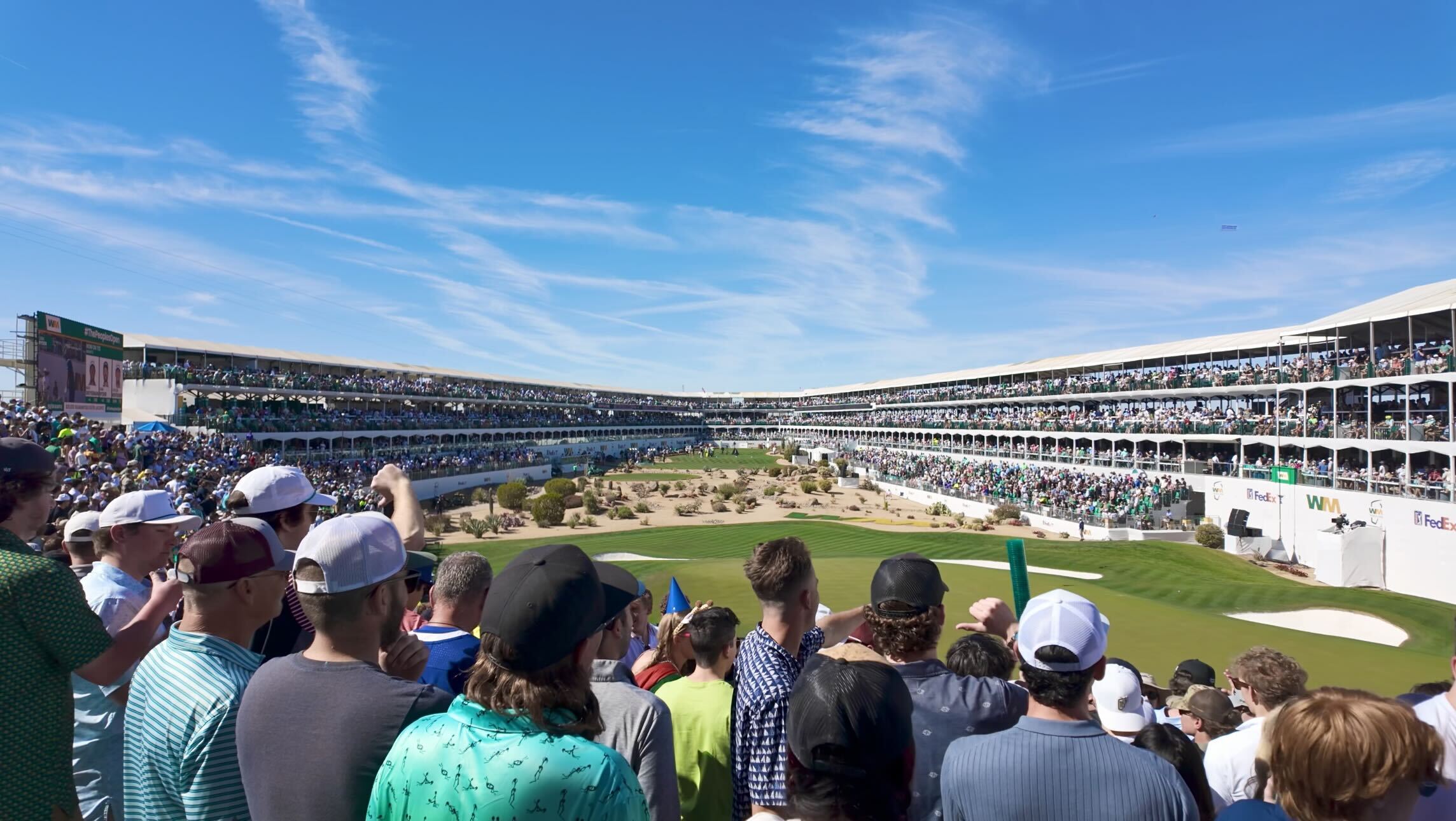 A large crowd gathers around a golf course with tiered seating, under a clear blue sky, watching a golf event.