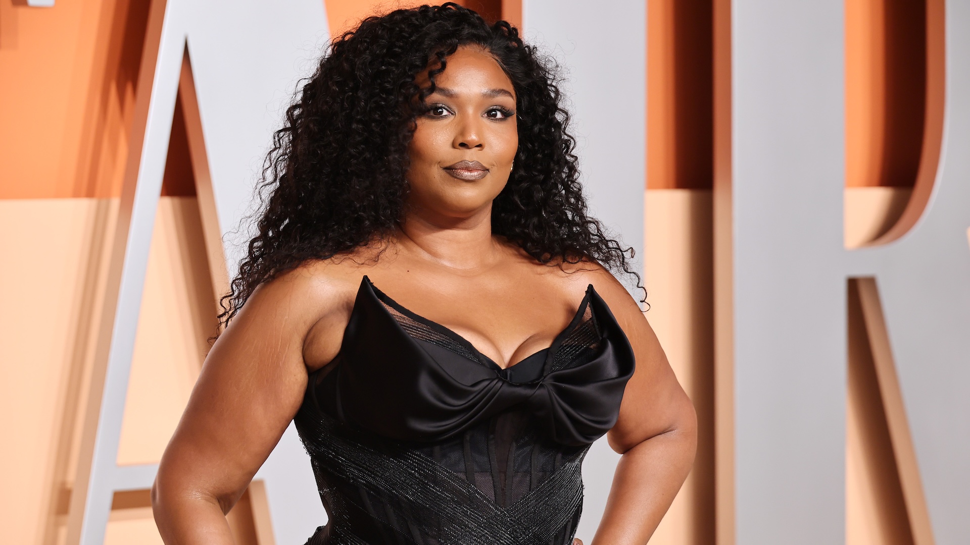 Lizzo poses in a black dress with a bow design at an event, with a backdrop of large letters.