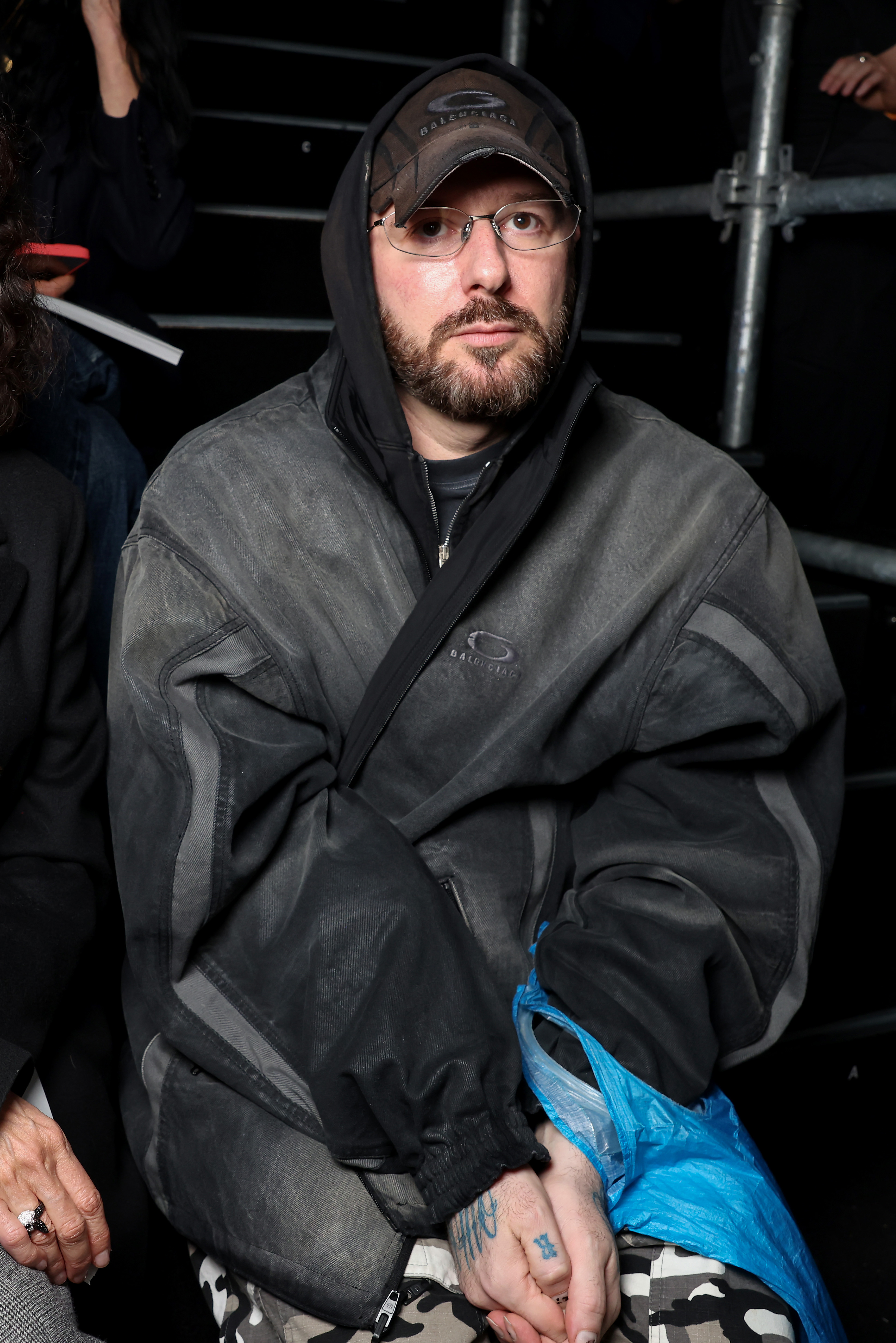 A man in a dark jacket and cap sits with hands clasped, wearing glasses, in an indoor setting.