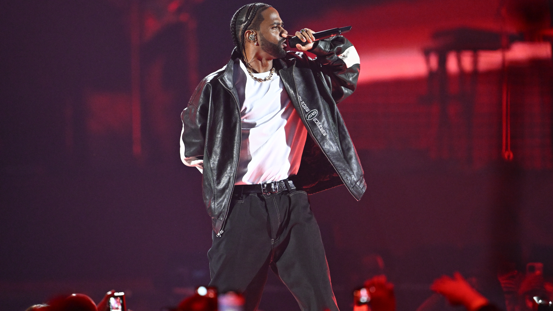 A performer in a leather jacket and white shirt sings on stage, holding a microphone, with a red-lit background and audience.