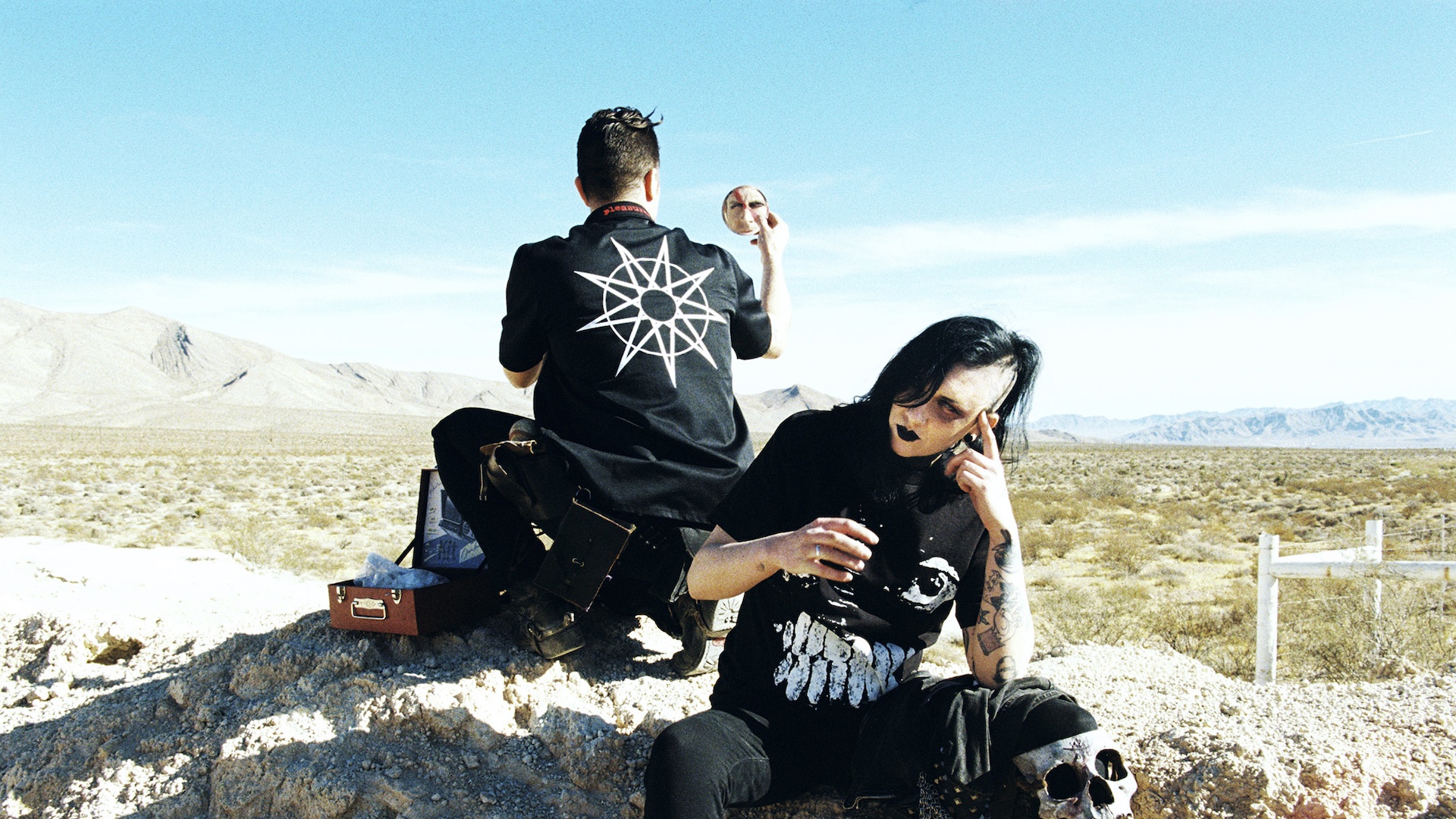 Two people in gothic attire sit in a desert landscape. One faces away with a star design on their shirt, the other with dark makeup.