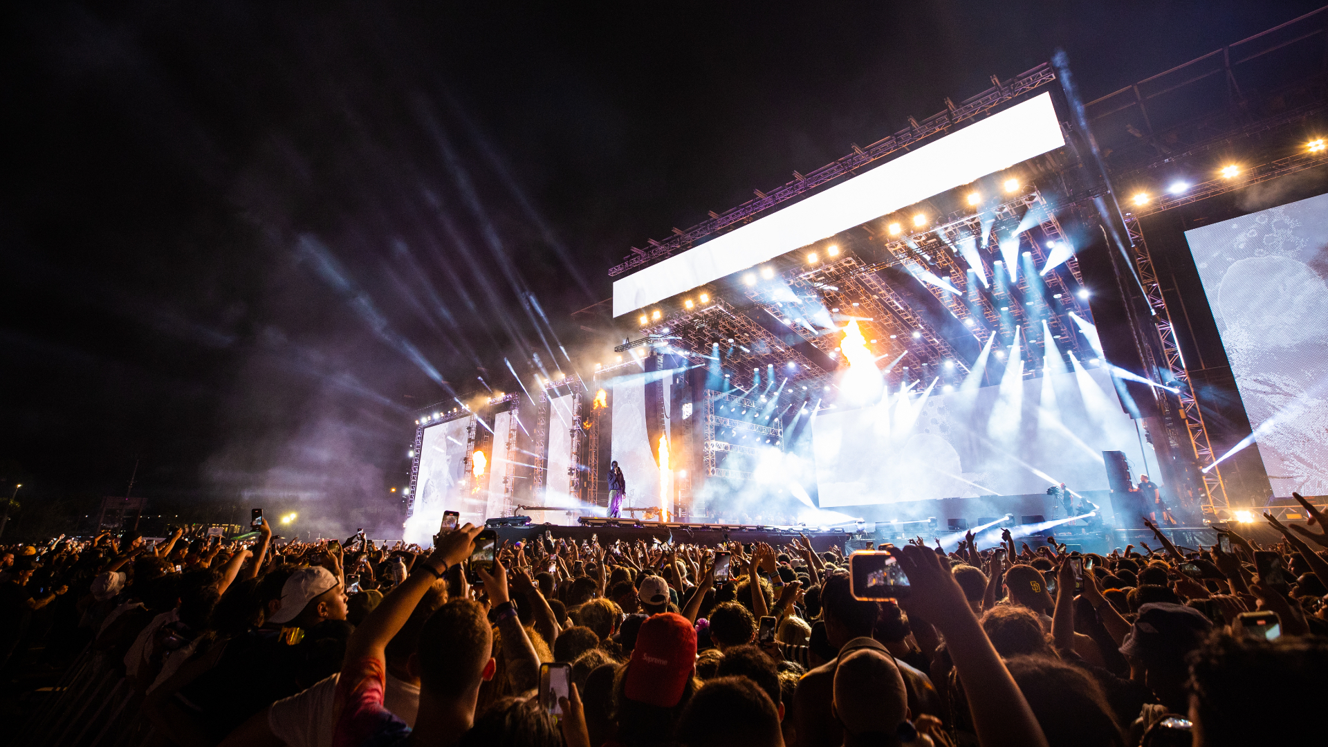 A large crowd at a nighttime concert with bright stage lights and a performer on stage. Fans are capturing the moment with phones.