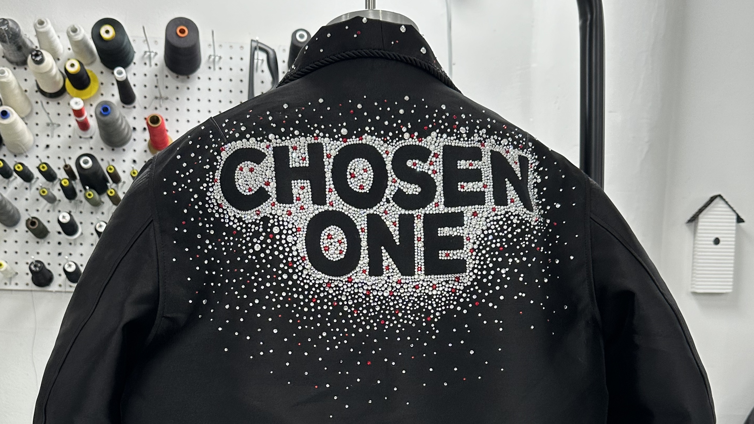 A black jacket with "CHOSEN ONE" in bold letters, adorned with a pattern of white and red dots. Sewing supplies are visible in the background.