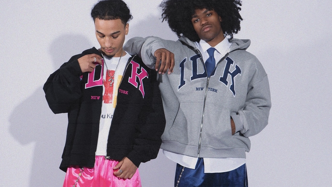 Two individuals wearing "IDK New York" hoodies, one in black with pink lettering, the other in gray with blue lettering. Casual fashion style.