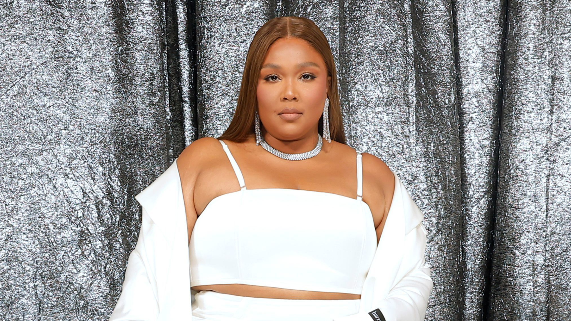 Lizzo in a white outfit, standing against a shimmering silver backdrop.
