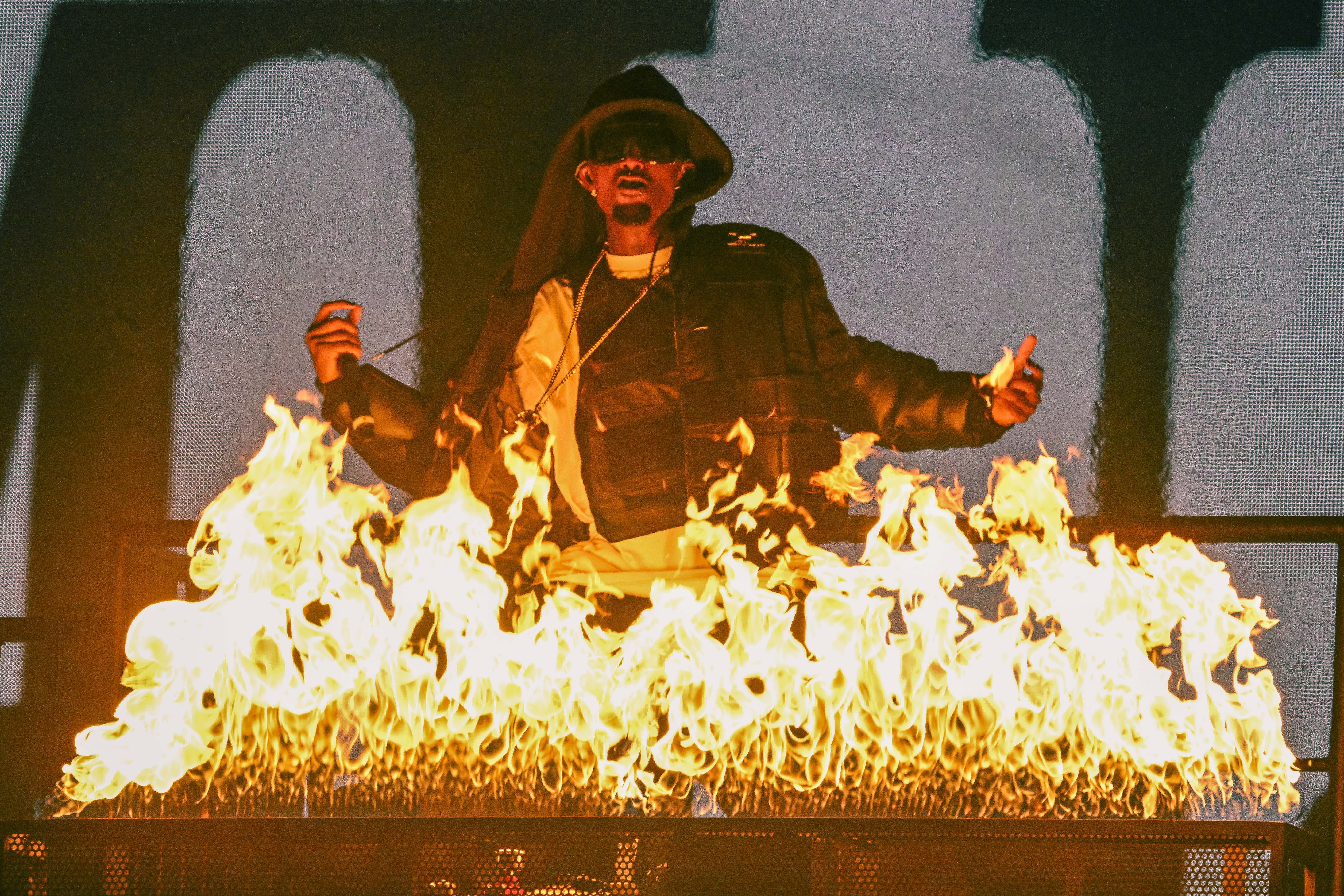 Playboi Carti on stage with a dramatic fire effect in front, wearing a hat and dark clothing, creating a dynamic and intense atmosphere.